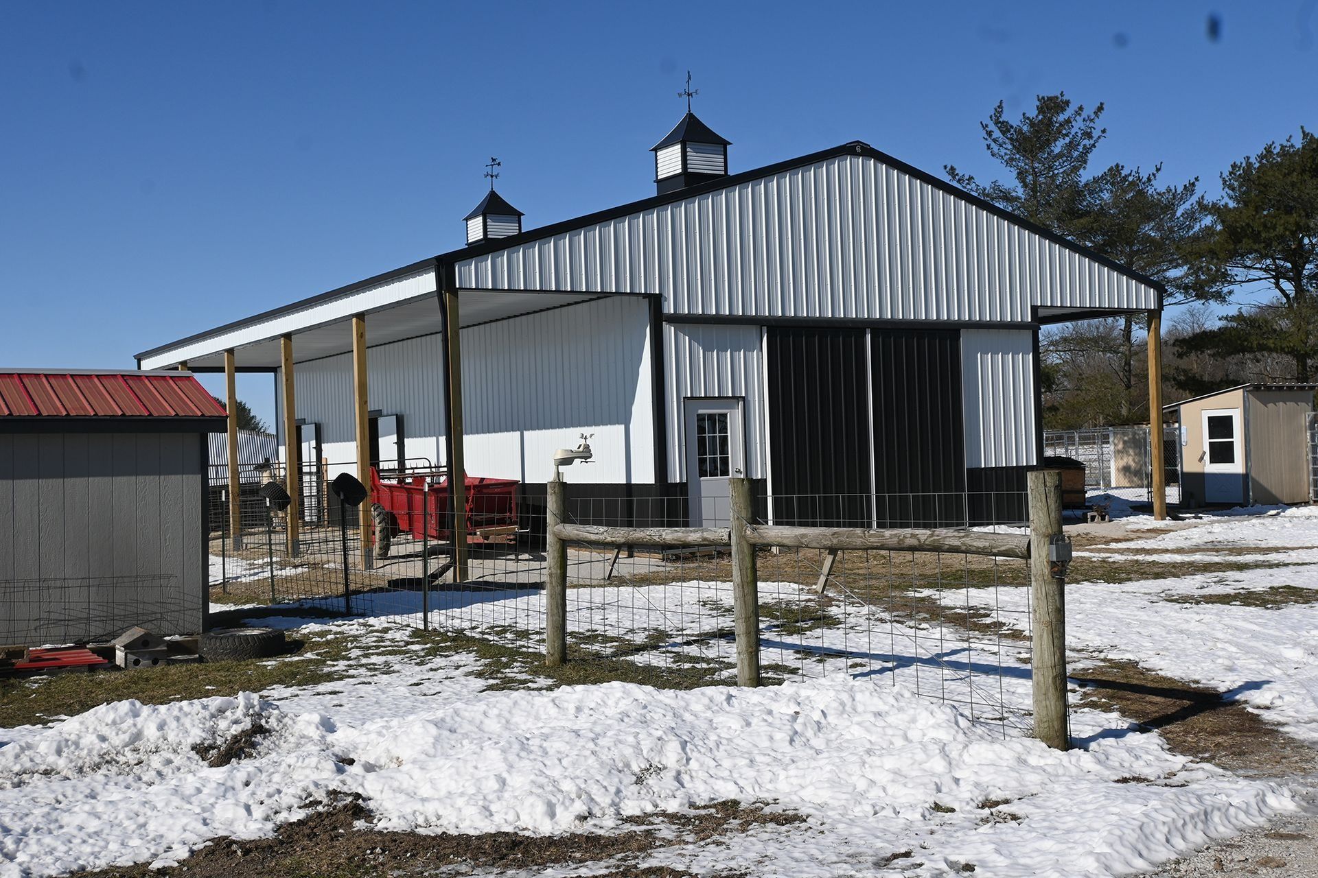 White metal barn with black accents, covered porch, snow on ground, blue sky.