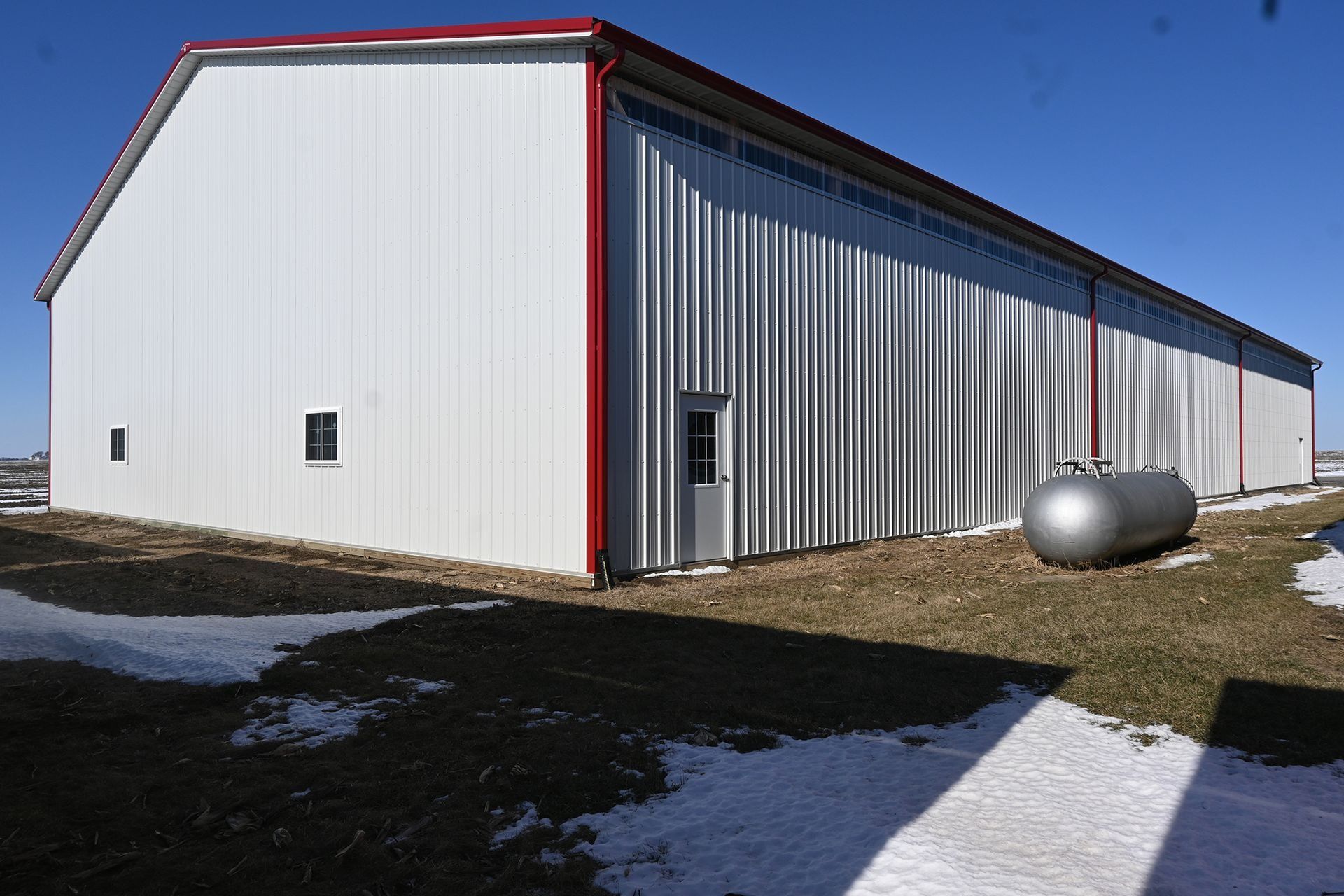 Large white metal building with red trim, door, and a propane tank outside, on a sunny day.