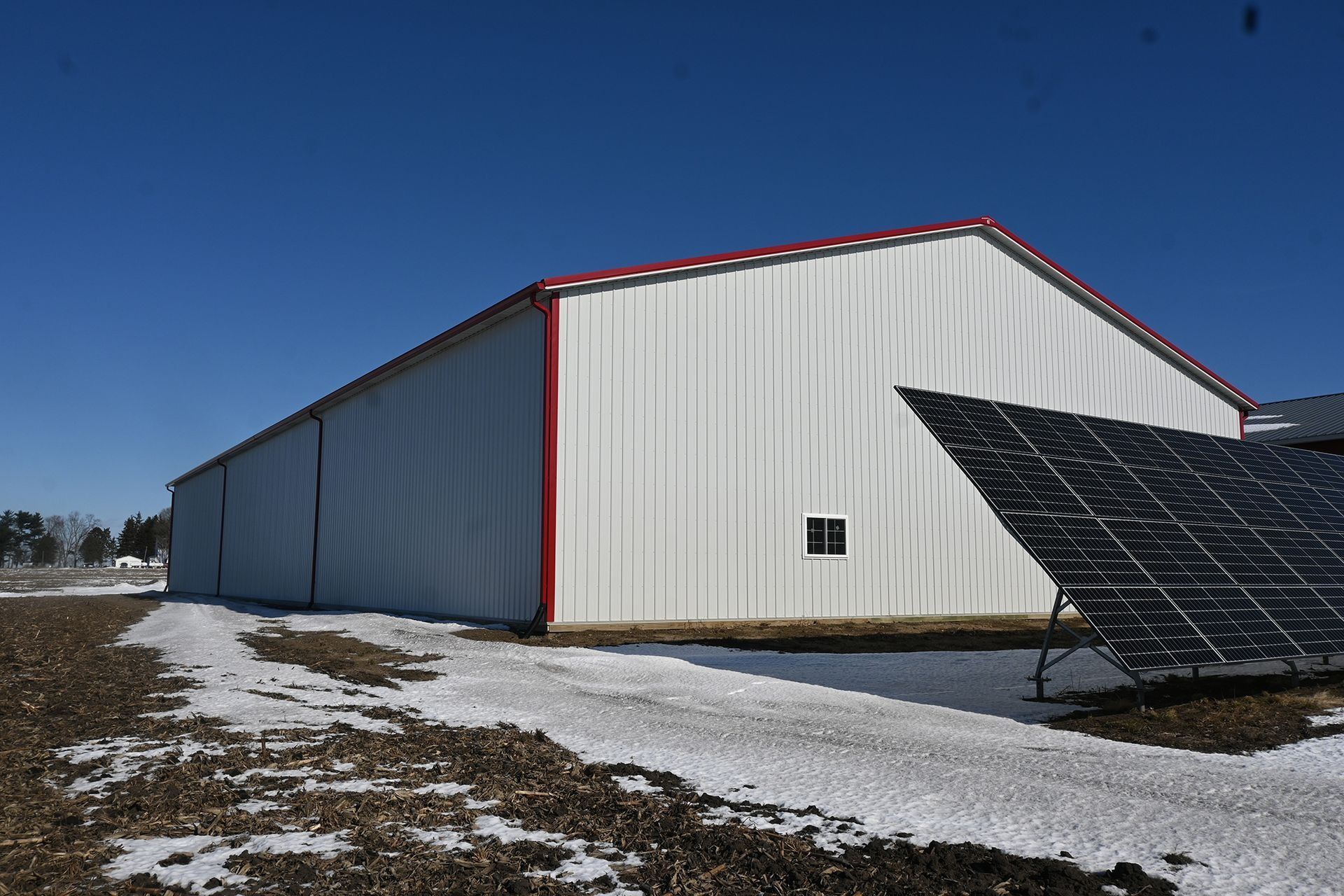 White metal barn with red trim, small window, and solar panels against a blue sky, snow on the ground.