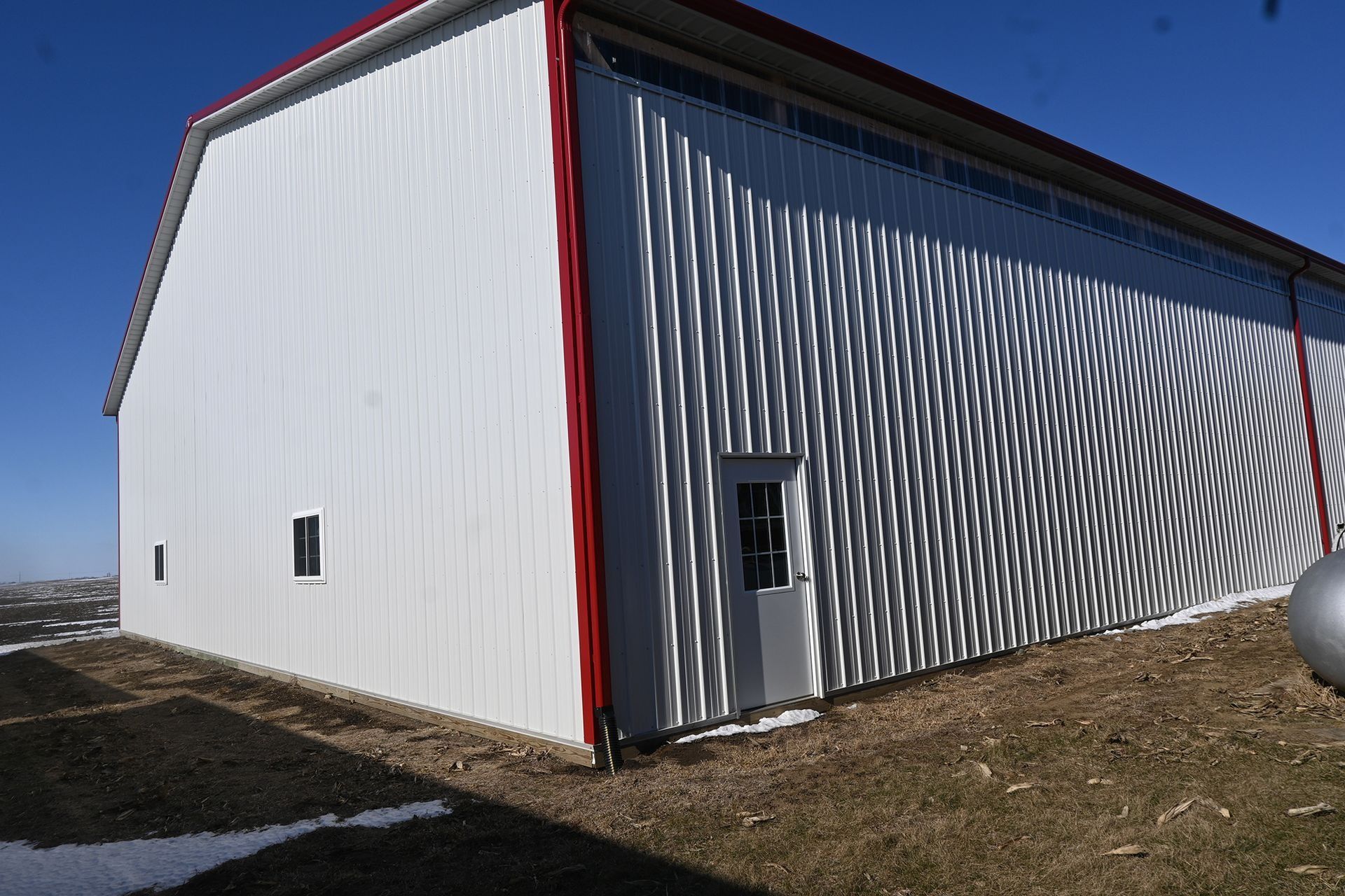 White metal barn with red trim, small windows, and a doorway, set against a blue sky.