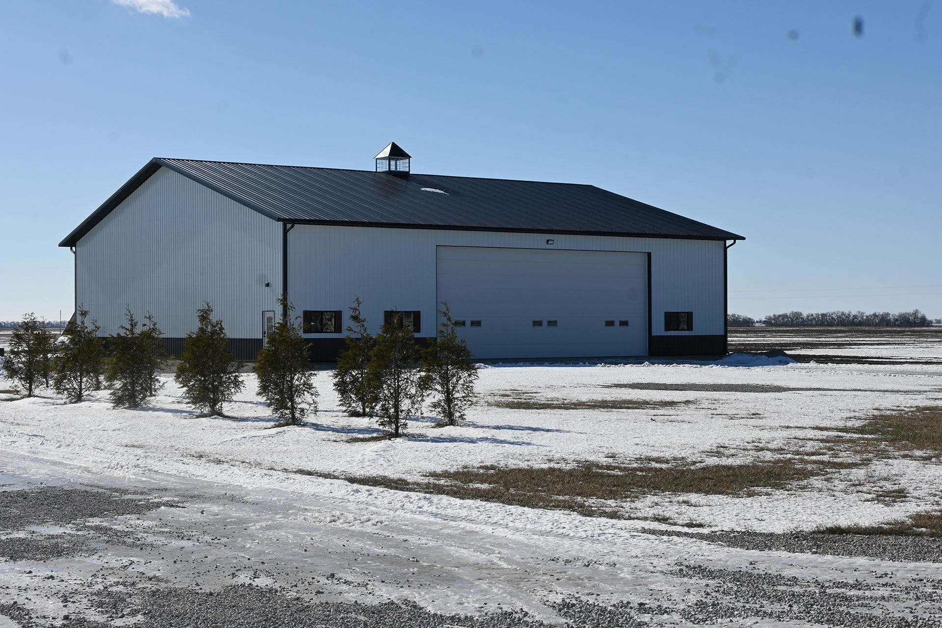 White metal building with a large garage door, set in a snowy field under a blue sky.