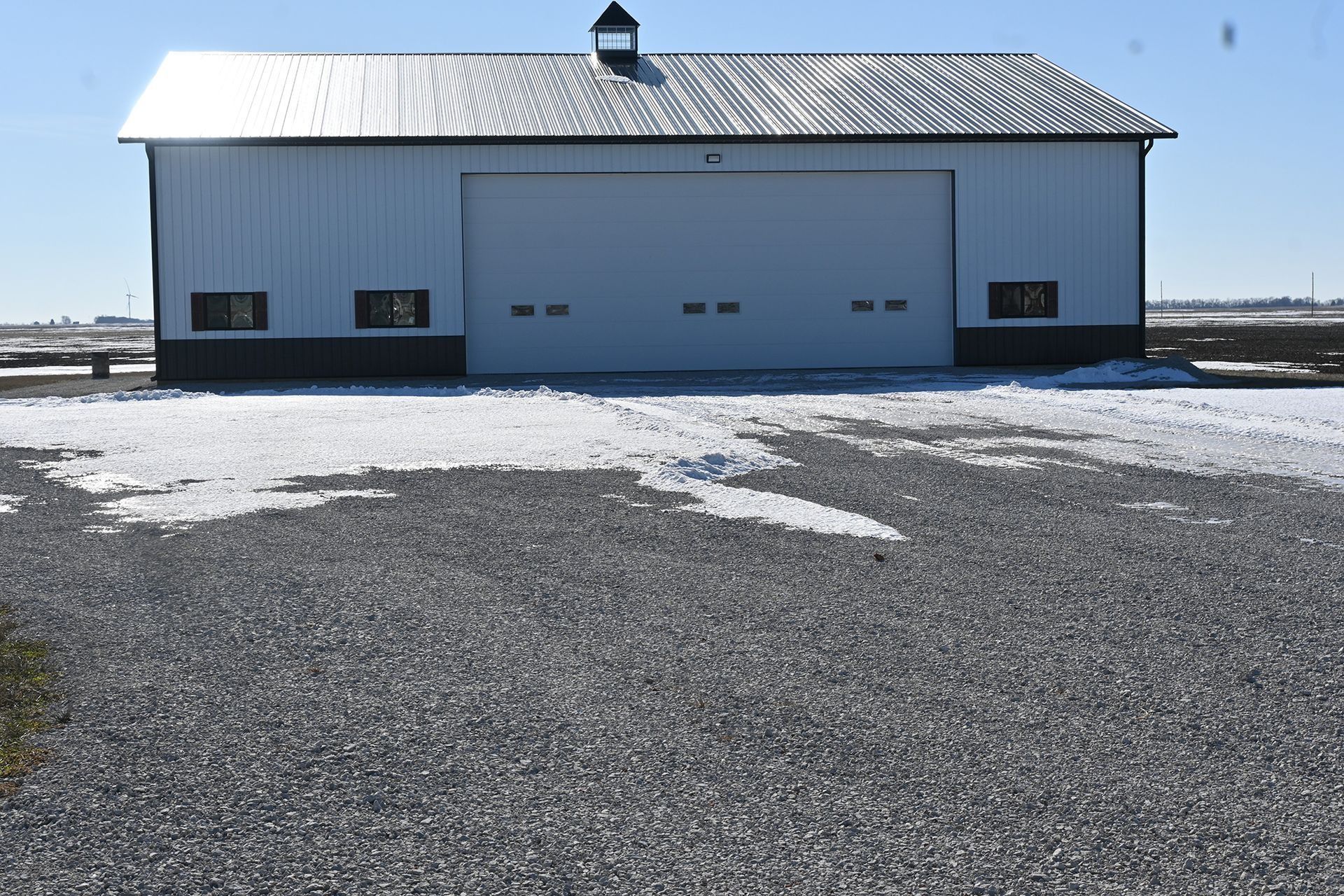 White and black barn with a large overhead door and gravel driveway, snow present.