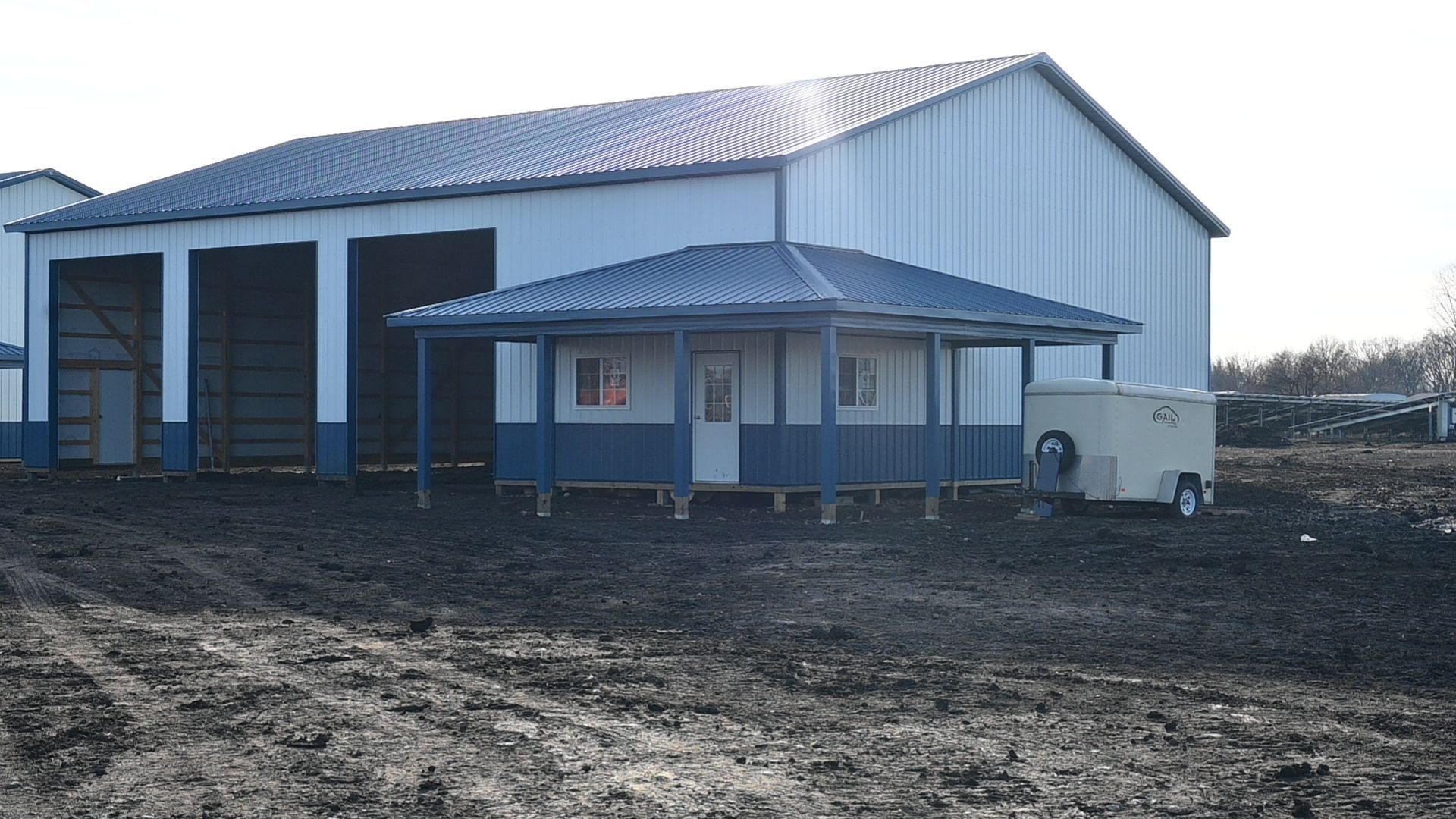 A small, blue and white building with a covered porch in front of a large metal warehouse.