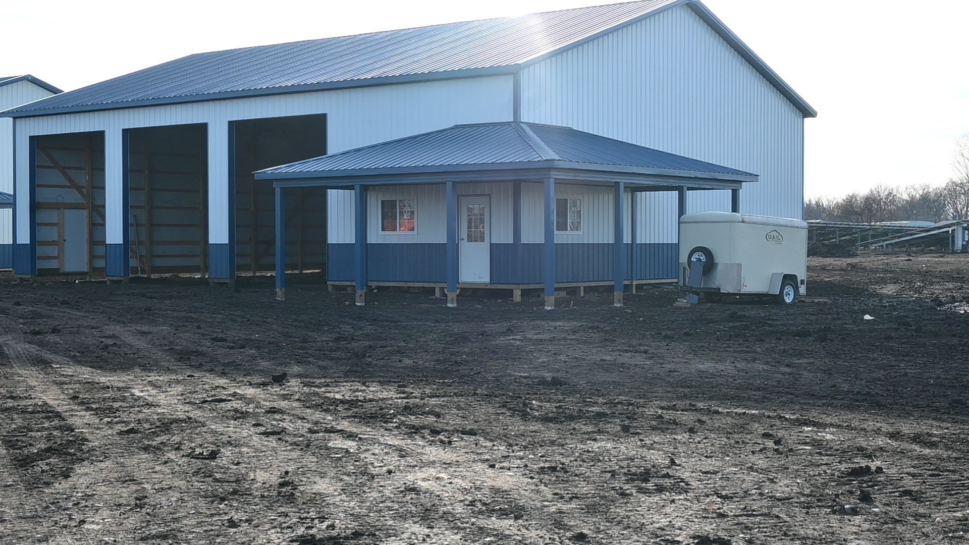 Small blue and white building with a porch in front of a large barn; muddy ground.