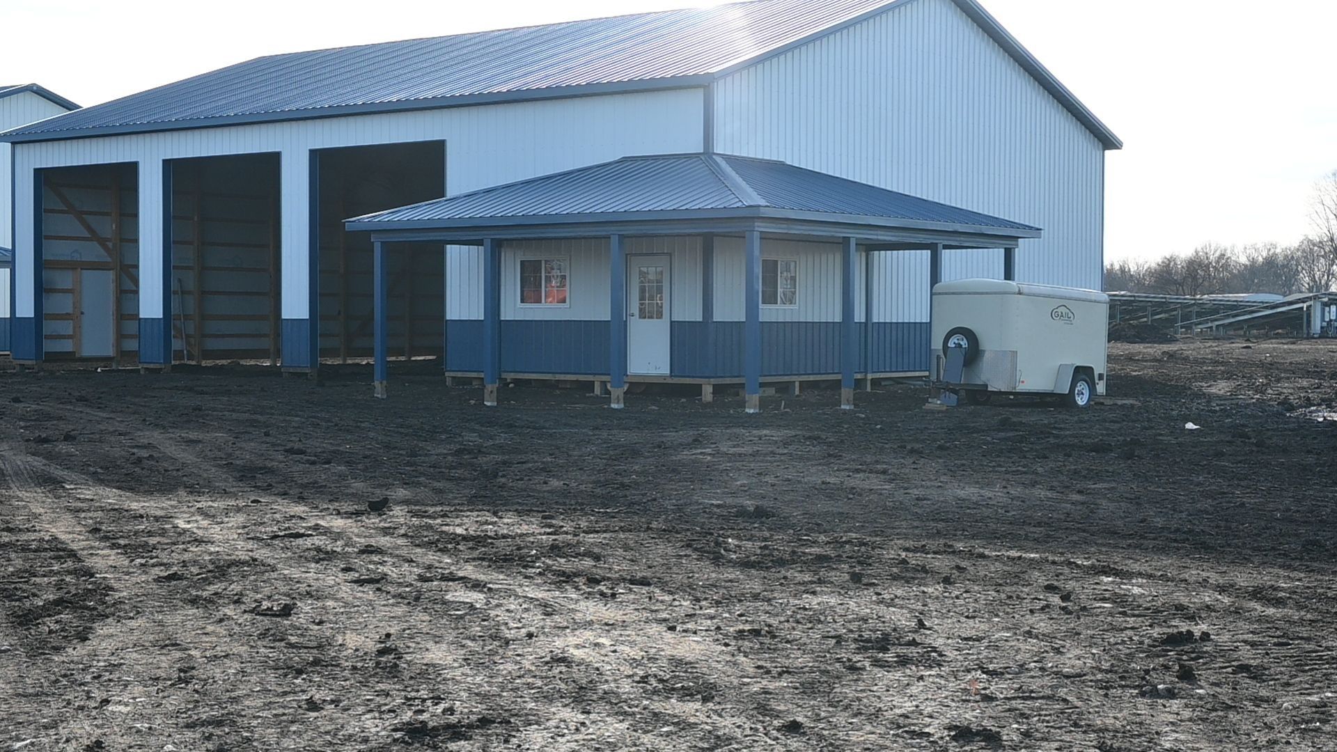 Small blue and white building with a porch, next to a large white barn and a trailer on muddy ground.