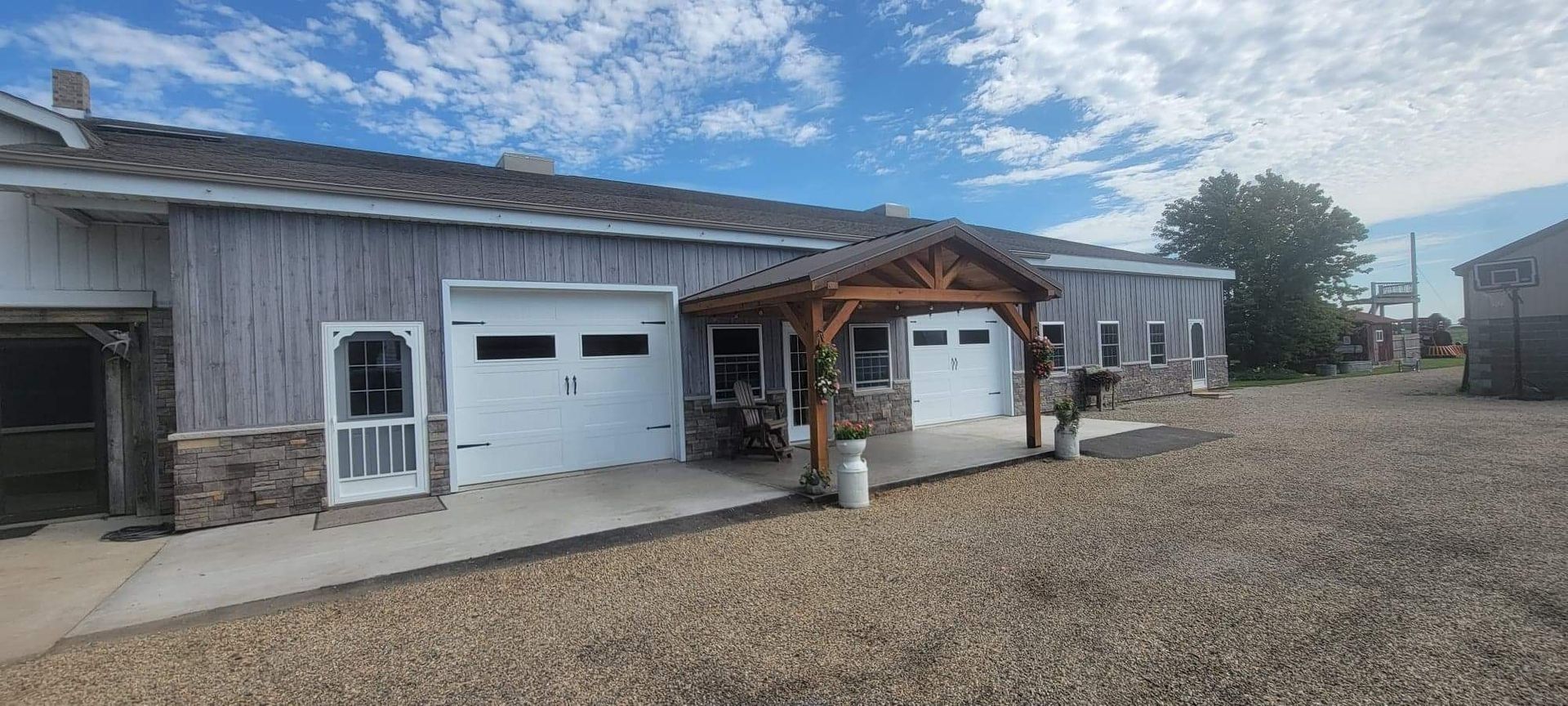 A gray barn with white garage doors and a wooden entryway. Gravel driveway under a blue sky.
