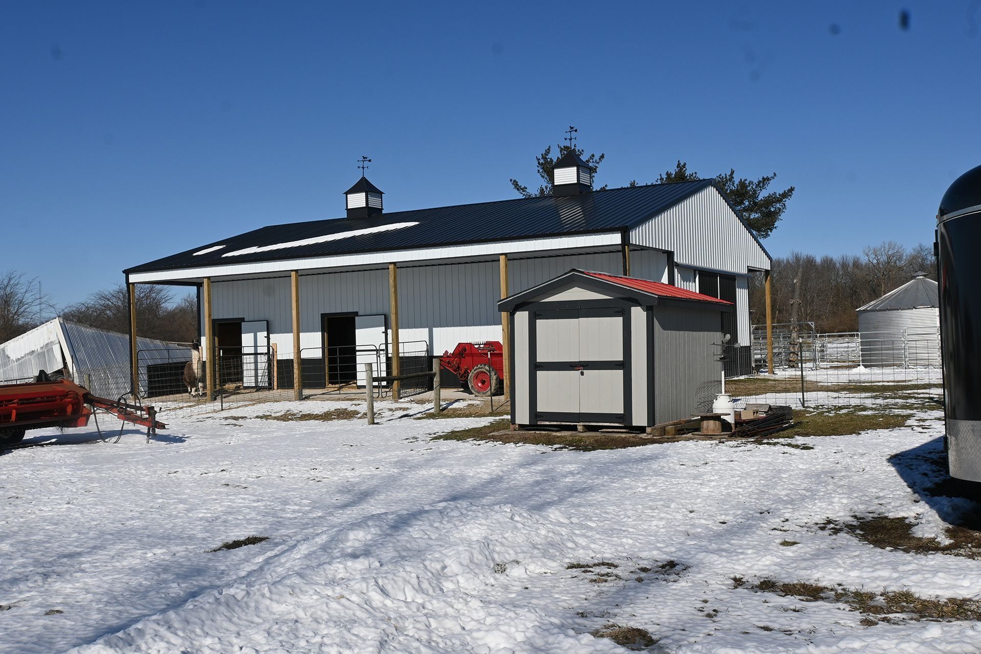 White barn with black roof, shed, and snow-covered ground on a sunny day.