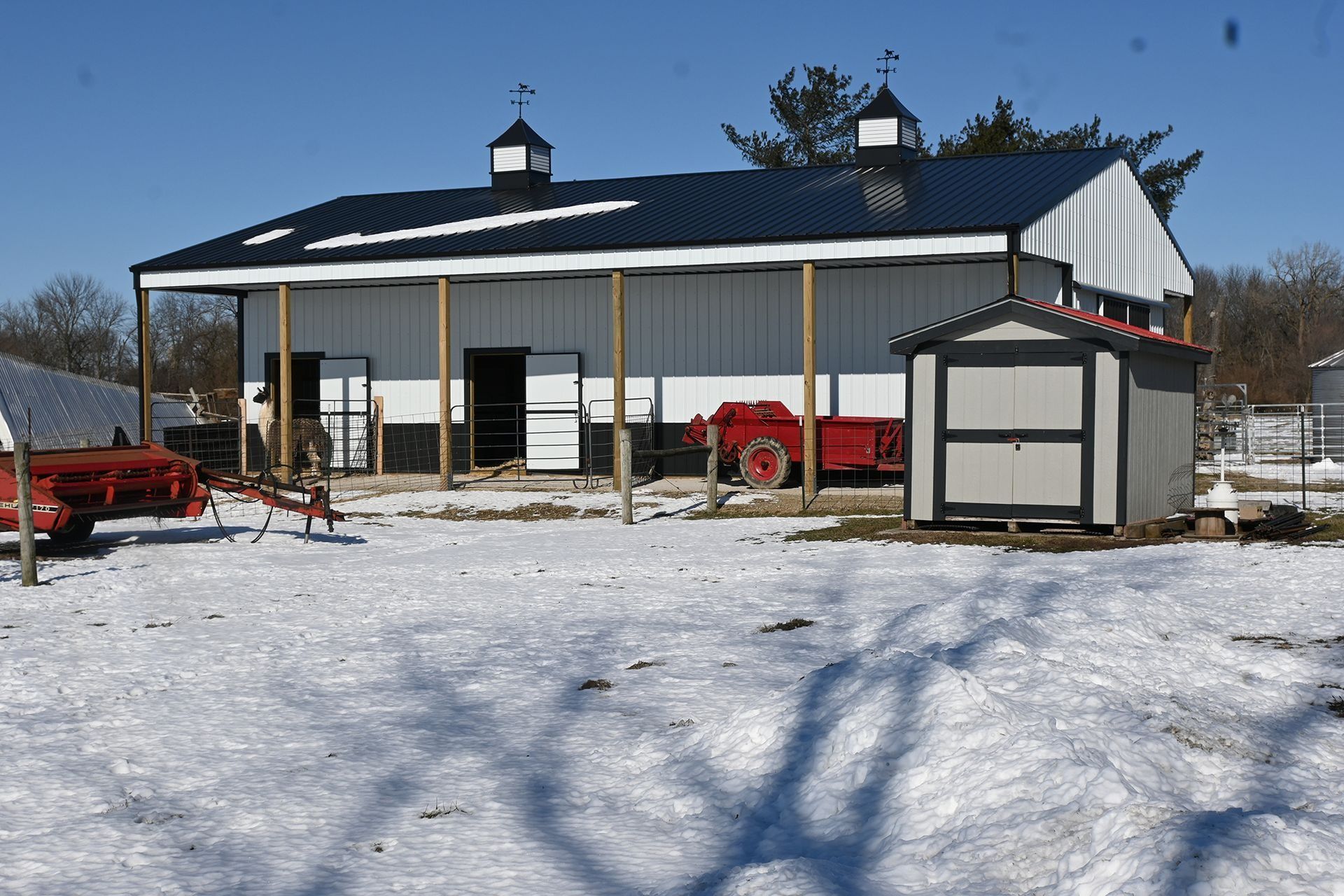 A white and black barn with a black roof, small shed, and tractor, set in a snowy field.