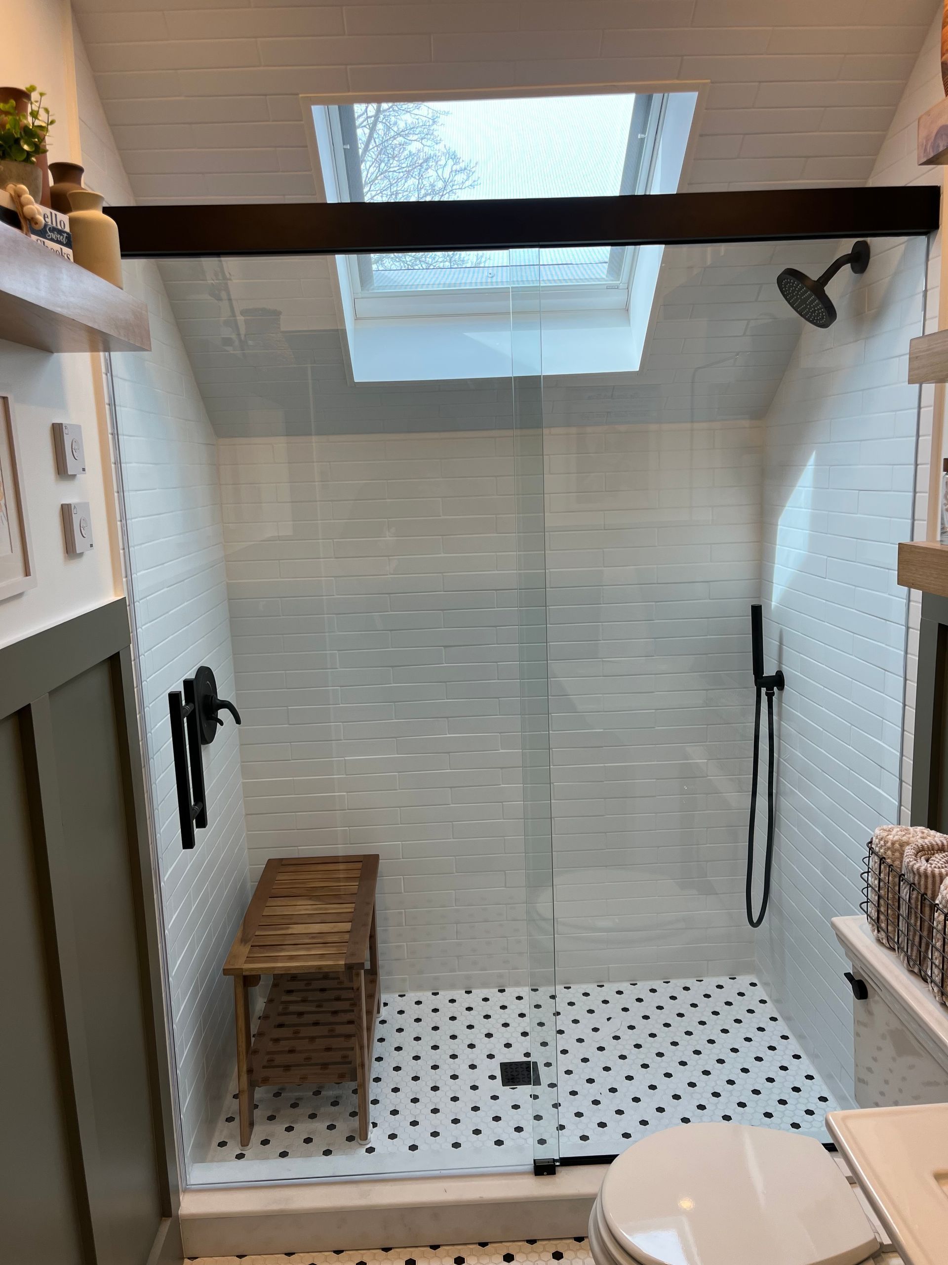 A modern walk-in shower with white-tiled walls, black fixtures, a patterned floor, a wooden bench, and a skylight above.