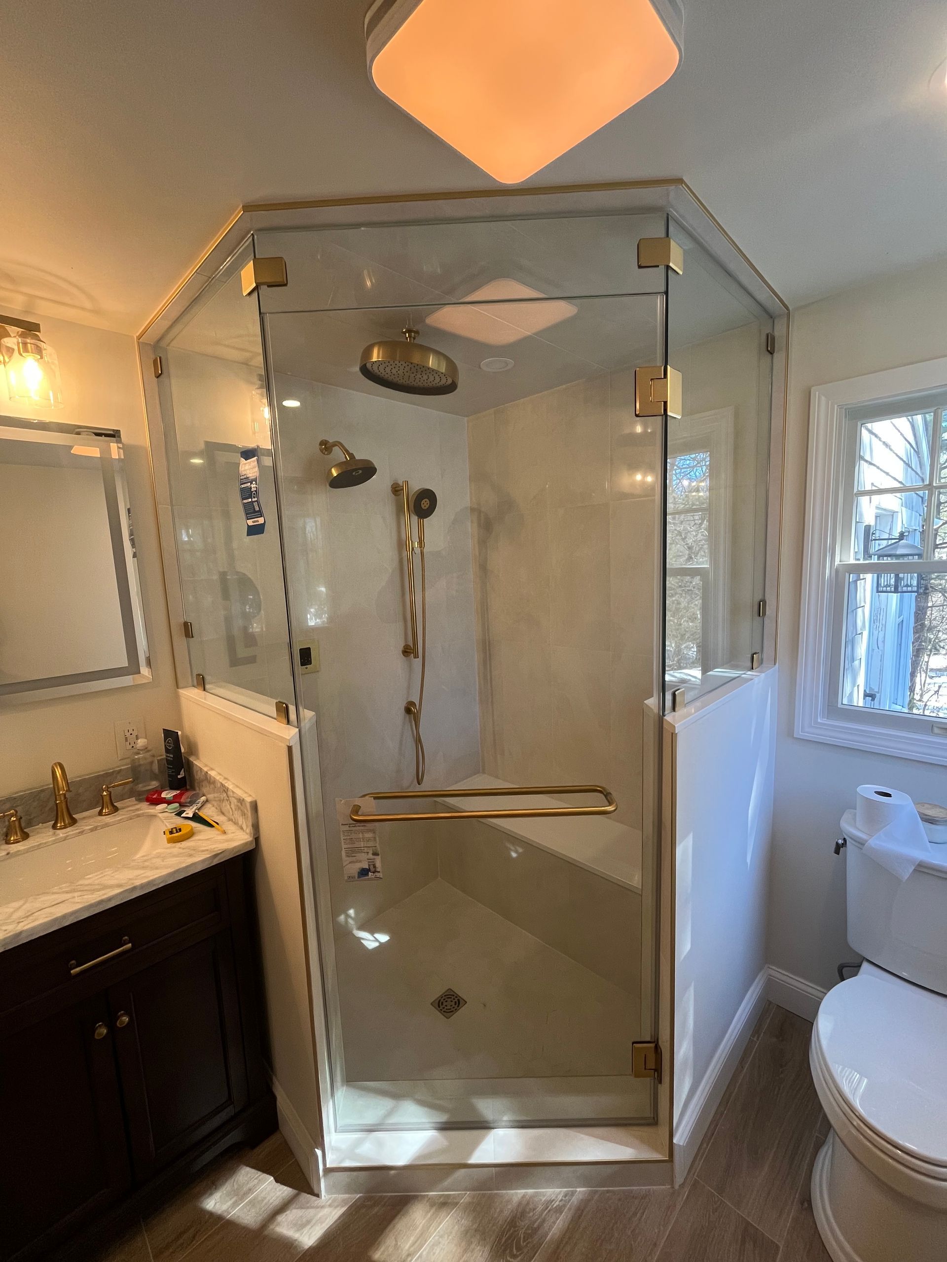 A modern bathroom with a glass-enclosed corner shower featuring gold fixtures, next to a dark vanity and a white toilet.