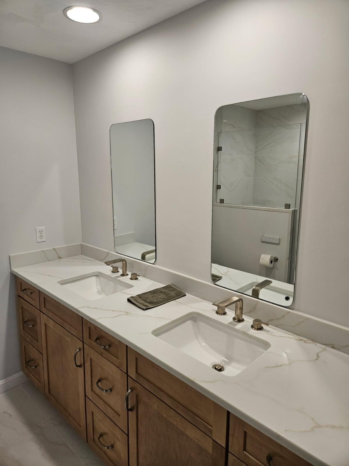 Bathroom vanity with double sinks, two mirrors, and light wood cabinets against a white wall.