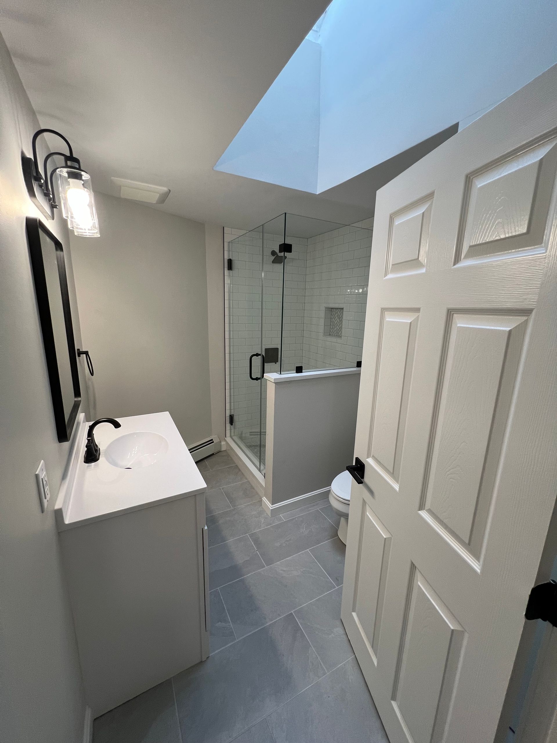 Modern bathroom with white vanity, glass shower, and skylight. Gray tiled floor and walls.