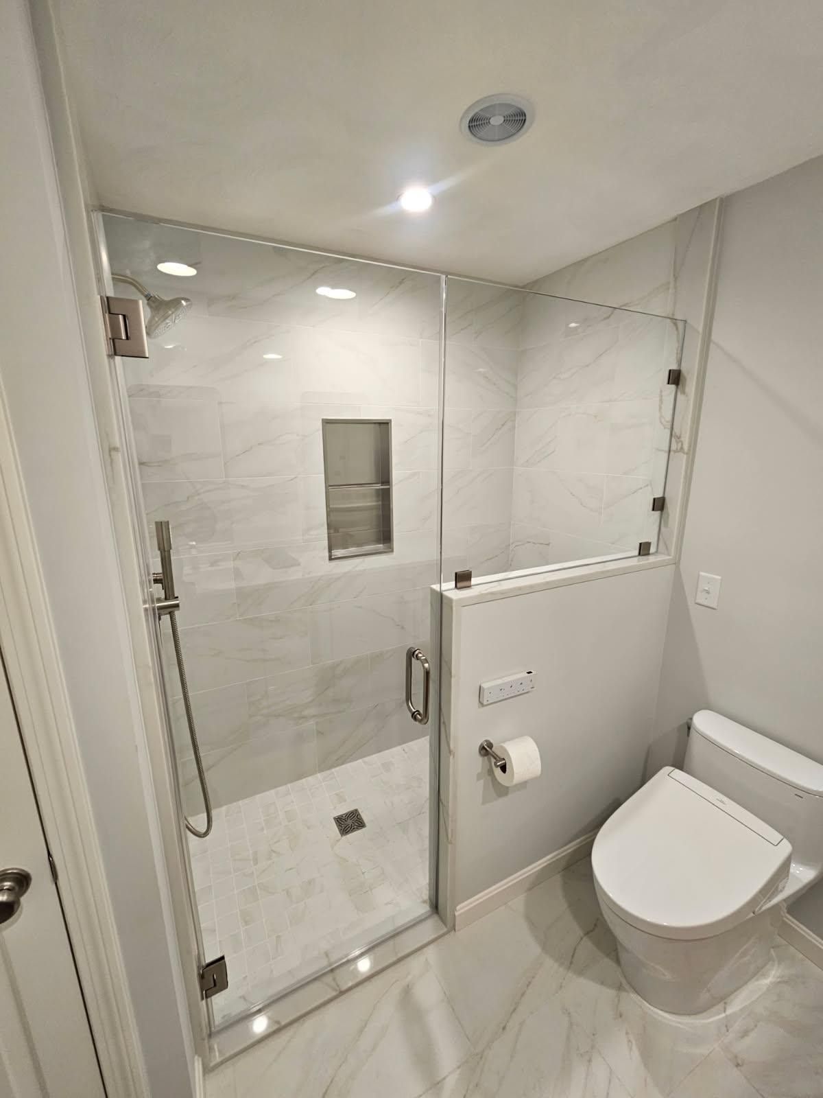 Glass-enclosed shower with white marble tile, toilet, and a recessed shelf.