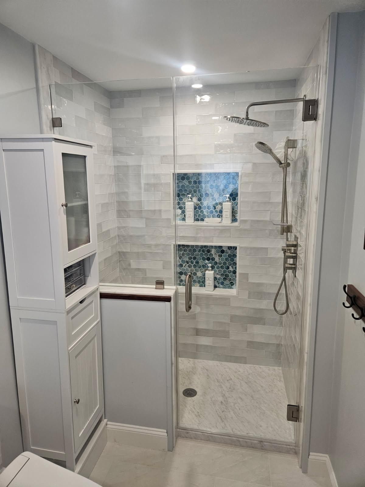 Modern bathroom with a glass shower, white cabinets, and gray tiled walls.