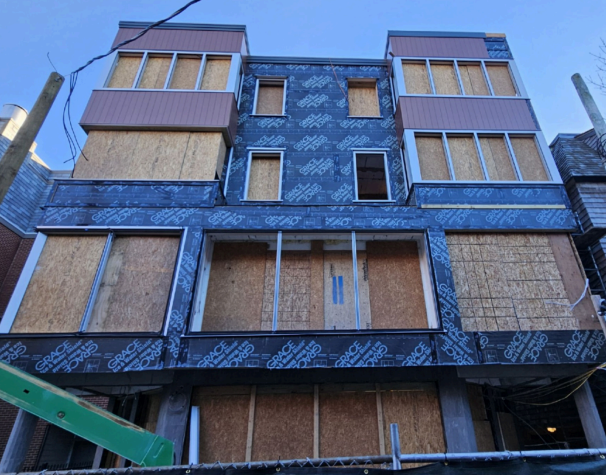 Building under construction, front view. Boarded up windows, dark siding, brown panels.