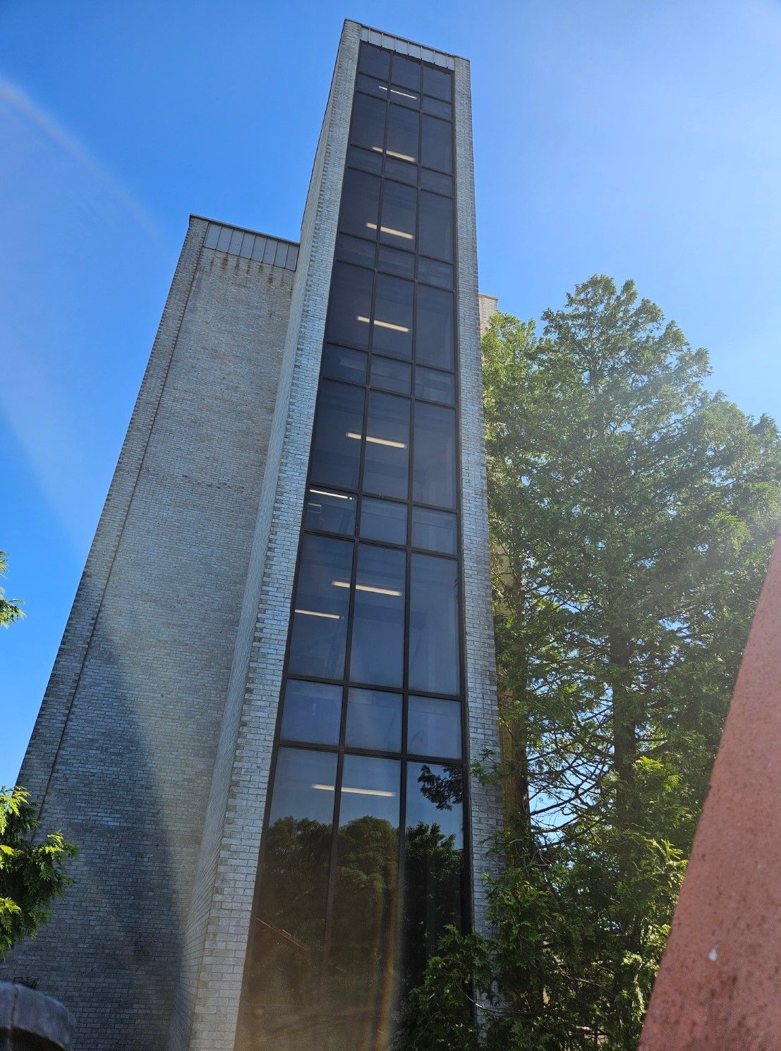 Tall brick building with a narrow, glass-windowed tower against a blue sky, trees on the right.