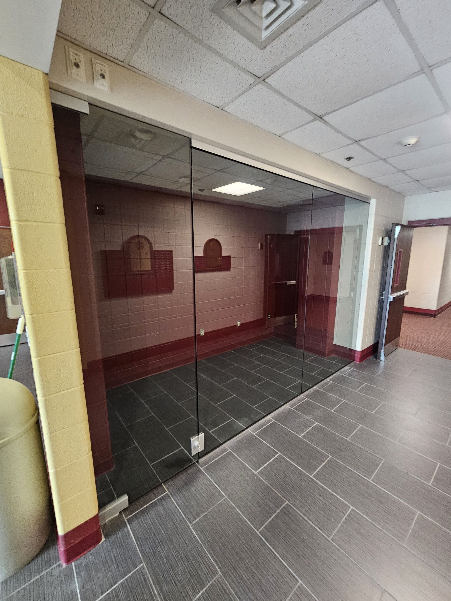Glass-walled room with dark tile floor, red accent wall, and beige column in a hallway with a drop ceiling.