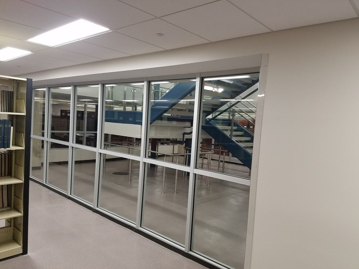 Library interior with glass windows and a staircase visible in the background. Beige walls and white shelving.