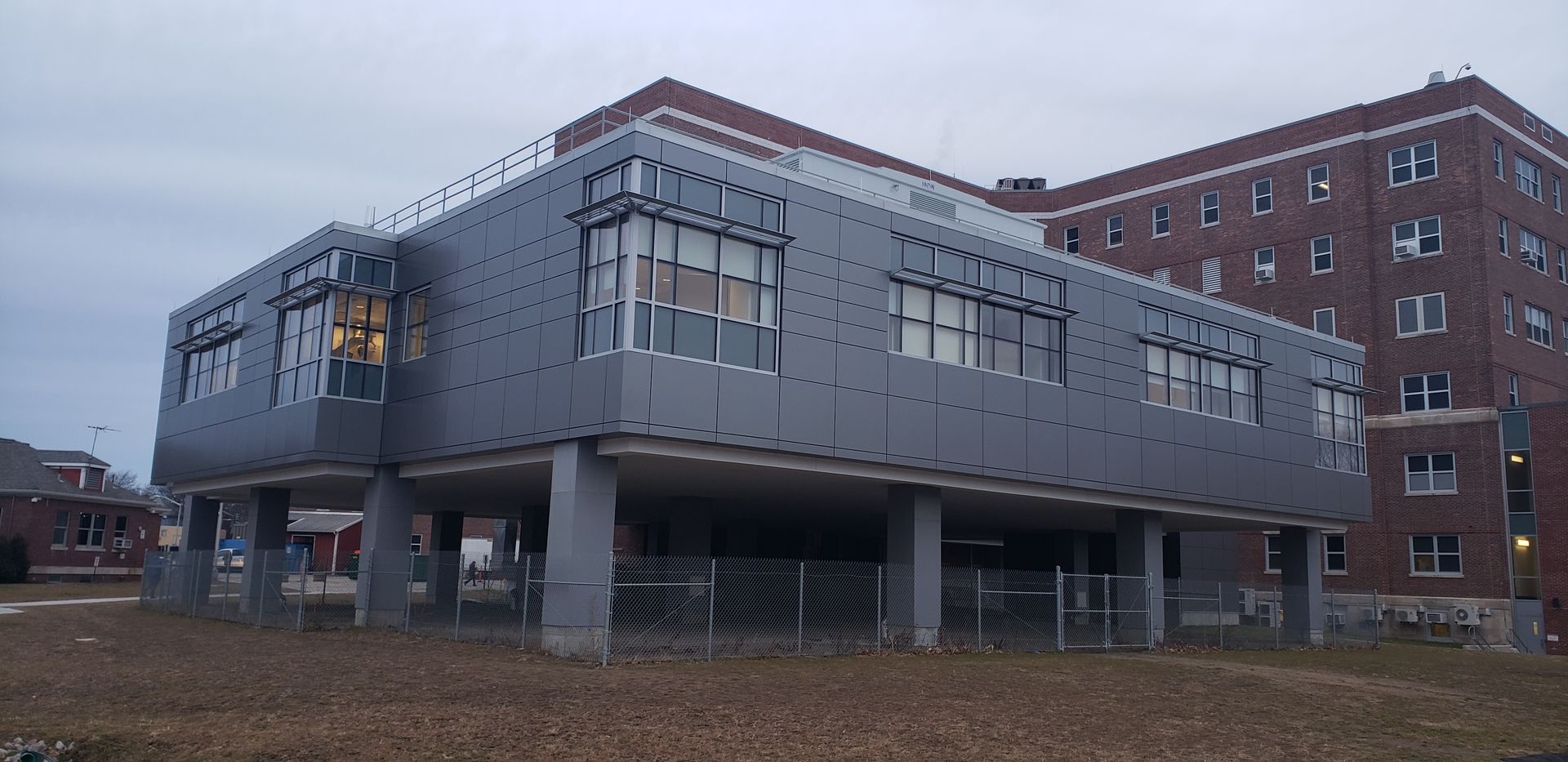 A modern building on stilts with a brick building in the background. Cloudy sky.