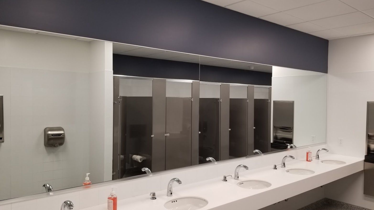 Public restroom with sinks, mirrors, and stalls. White walls, grey stalls, and blue accent.