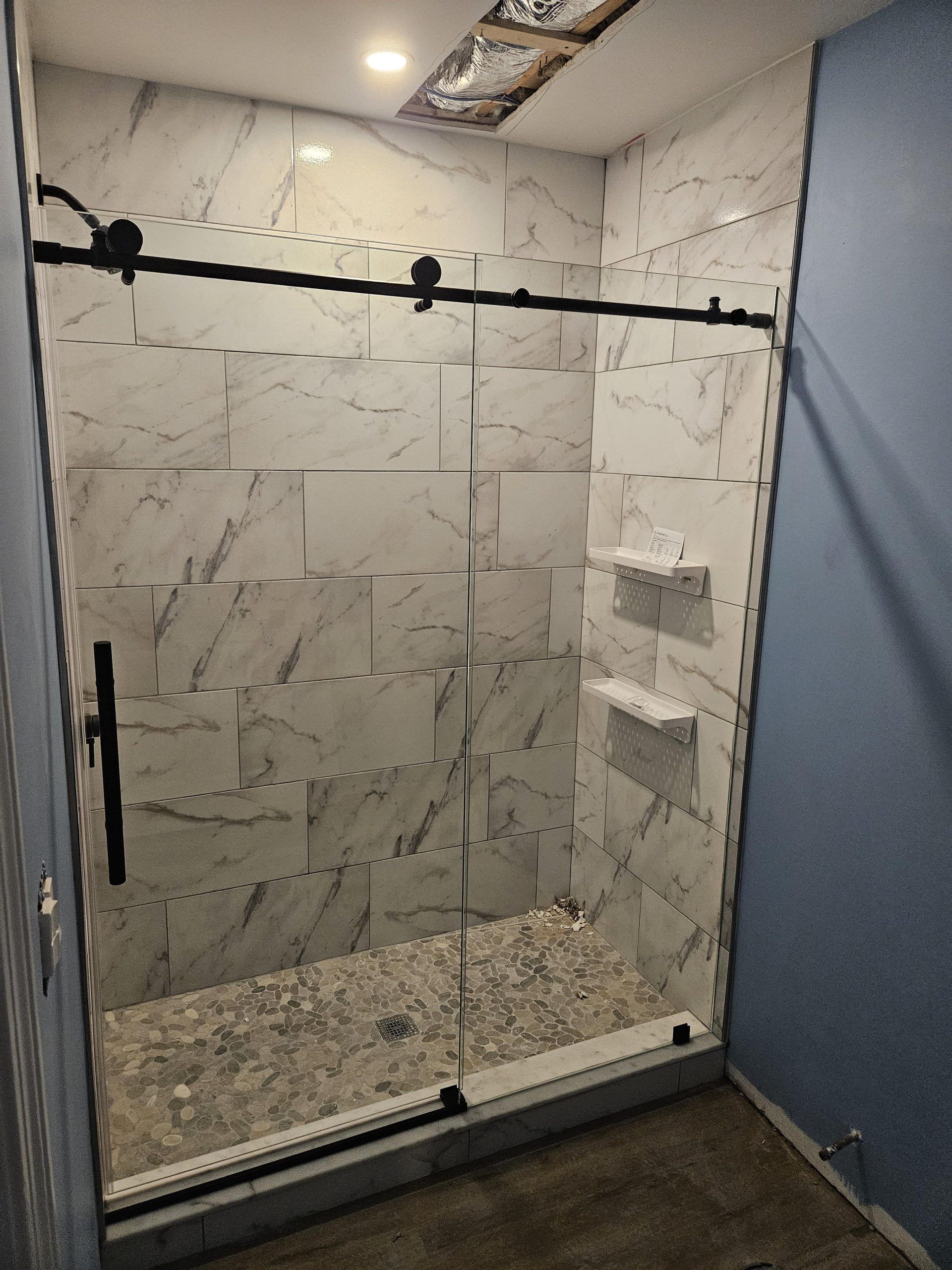 Shower with marble-look tile, glass door with black hardware, and pebble floor. Two built-in shelves.