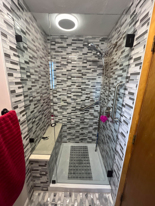Shower with gray, white, and black mosaic tile walls, glass doors, and a built-in bench.