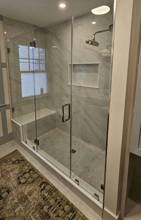 Glass-walled shower with white marble-look tiles, a built-in bench, and pebble-tile flooring, next to a window.