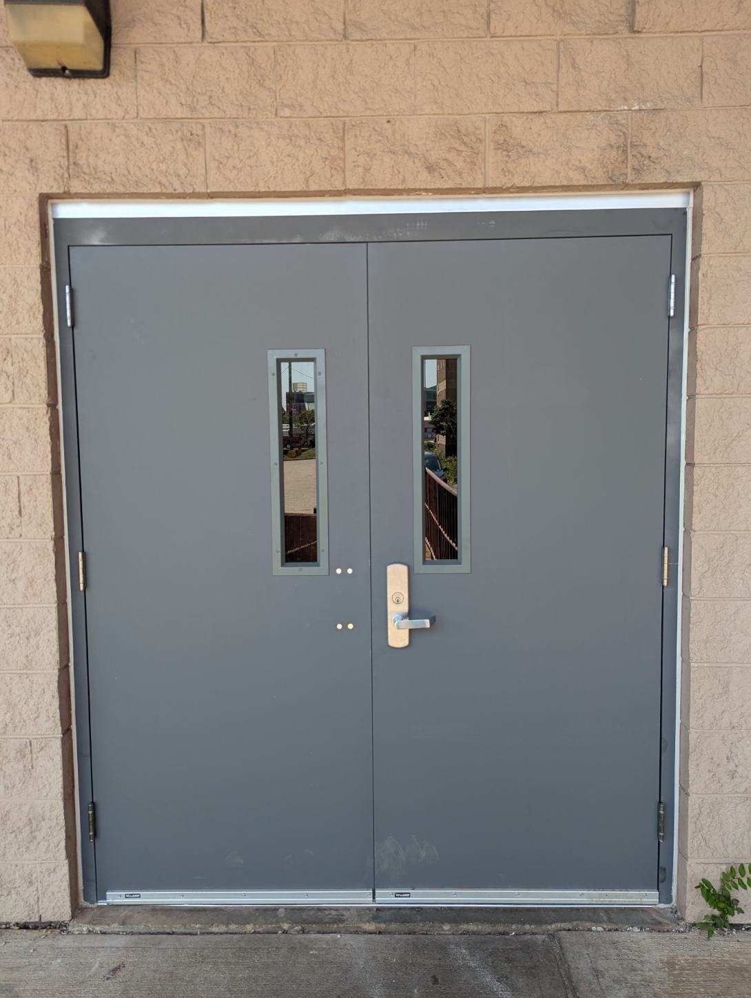 Gray double doors with two rectangular windows and a silver handle on a brick building.