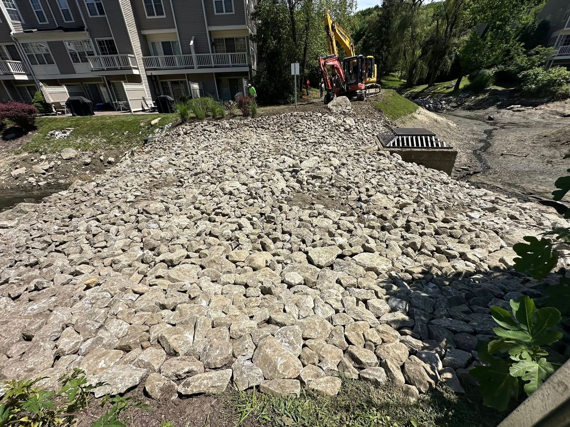 Rocky construction site with an excavator near a building and greenery.
