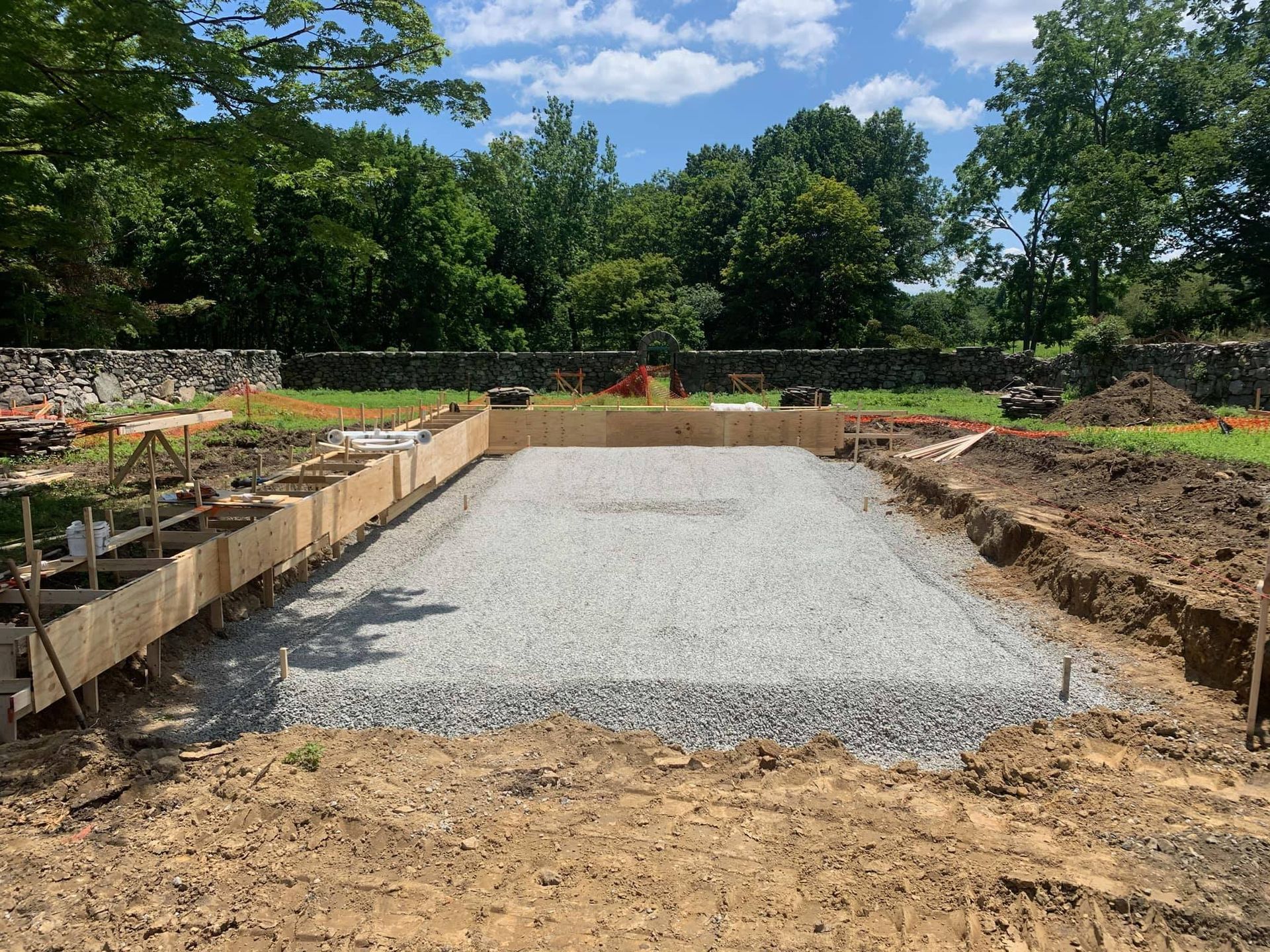 Construction site with gravel foundation and wooden forms, in a grassy area with trees and blue sky.