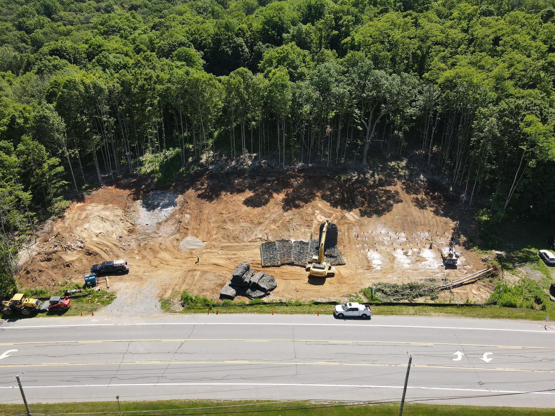 Construction site next to a highway. Excavators and trucks on dirt, trees in background, and cars driving.