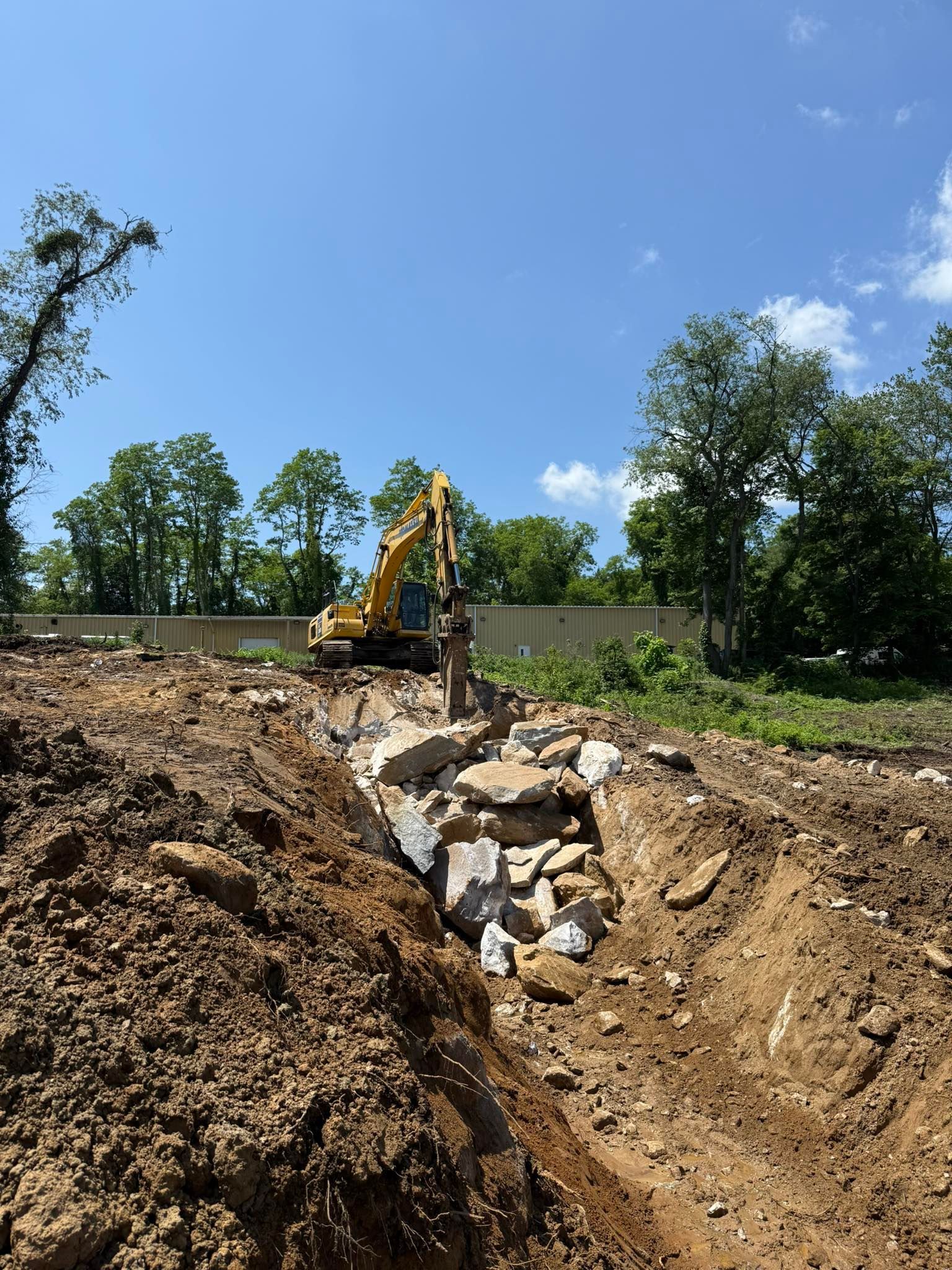 An excavator demolishes concrete amidst dirt, trees, and a blue sky.