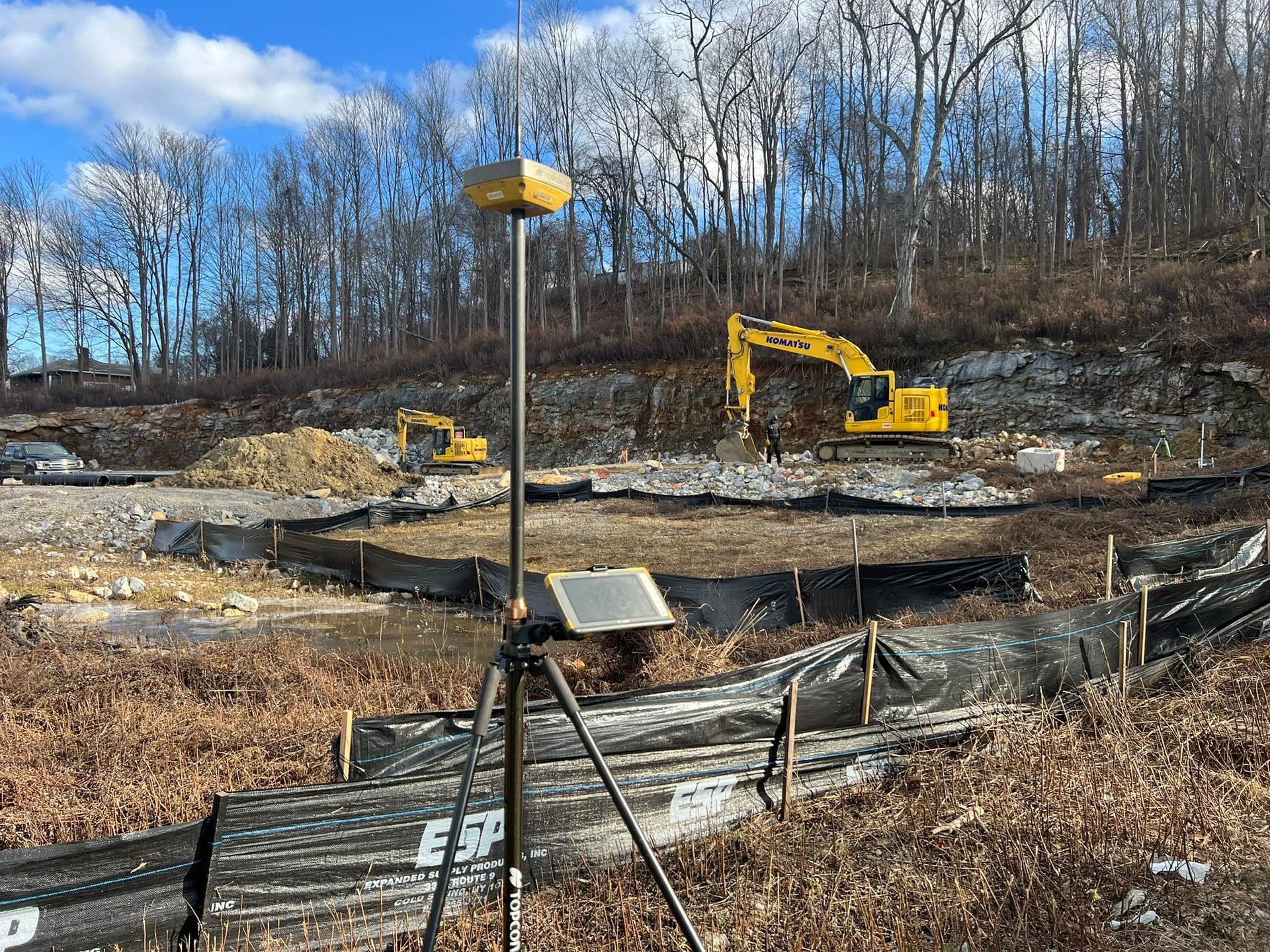 Construction site with GPS surveying equipment in the foreground, excavators, and wooded hillside.