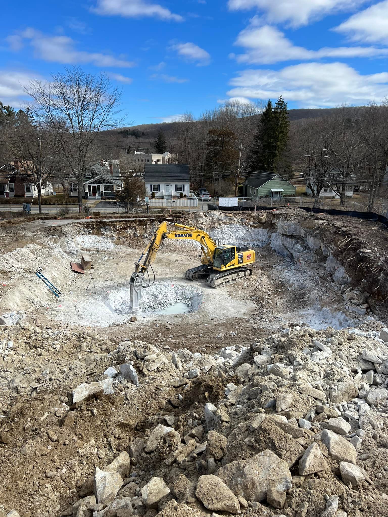 Yellow excavator in a large construction site. Cloudy sky, trees, and houses in the background.