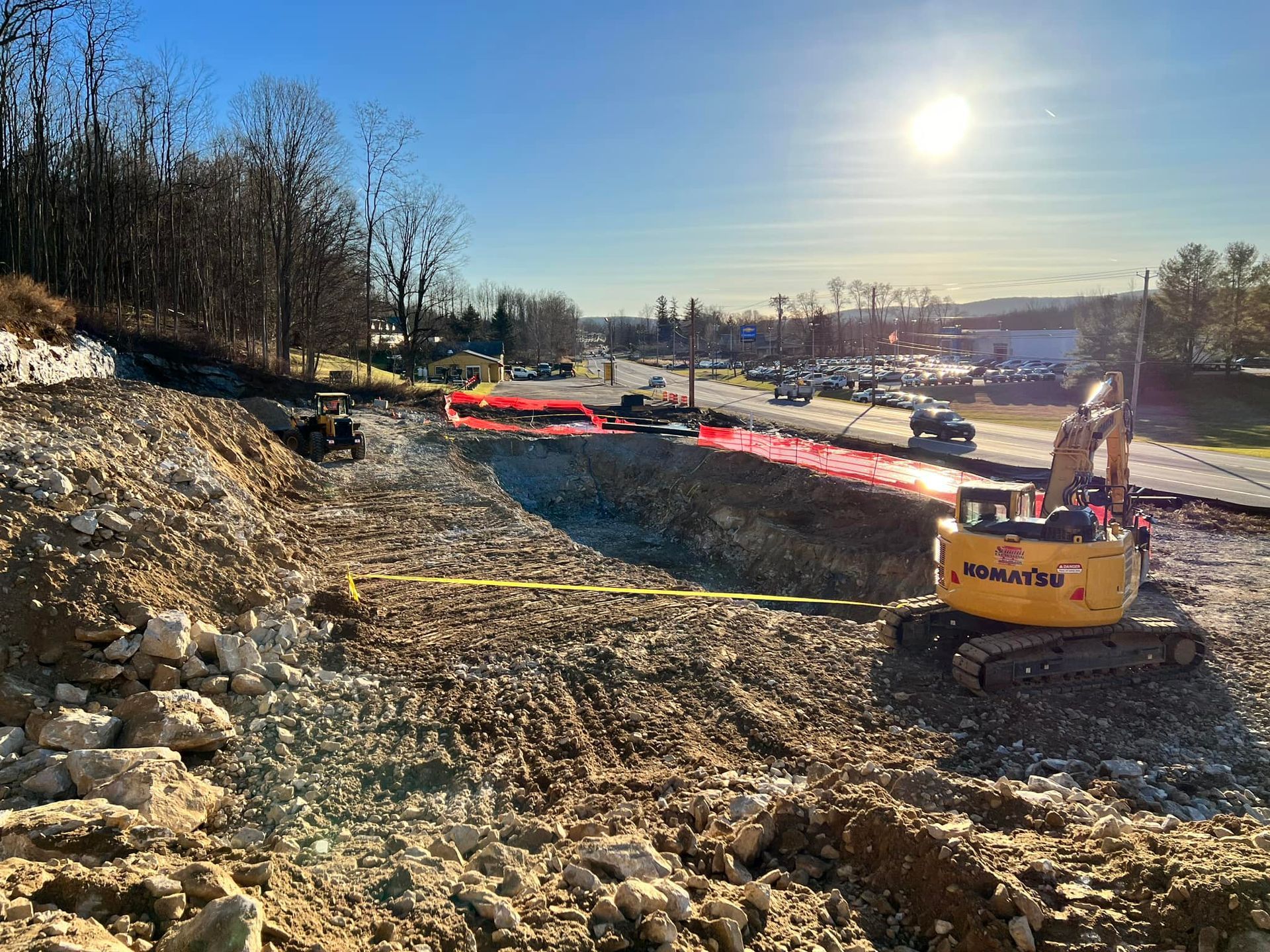 Construction site with an excavator. A road and trees are in the background. Bright sunlight.