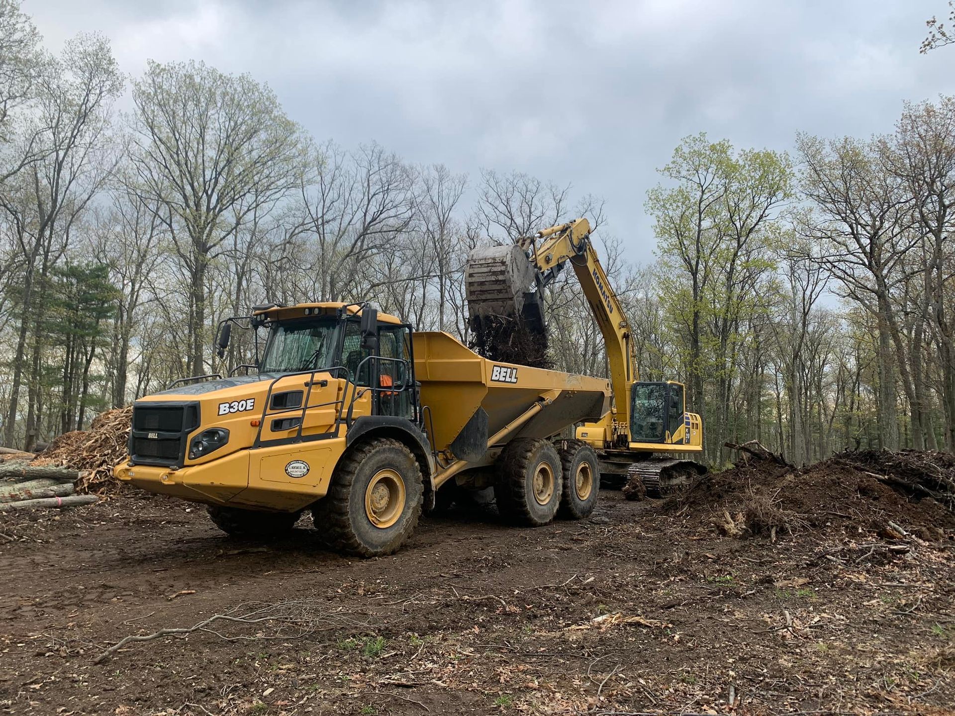 Yellow excavator loading a yellow dump truck in a wooded area.