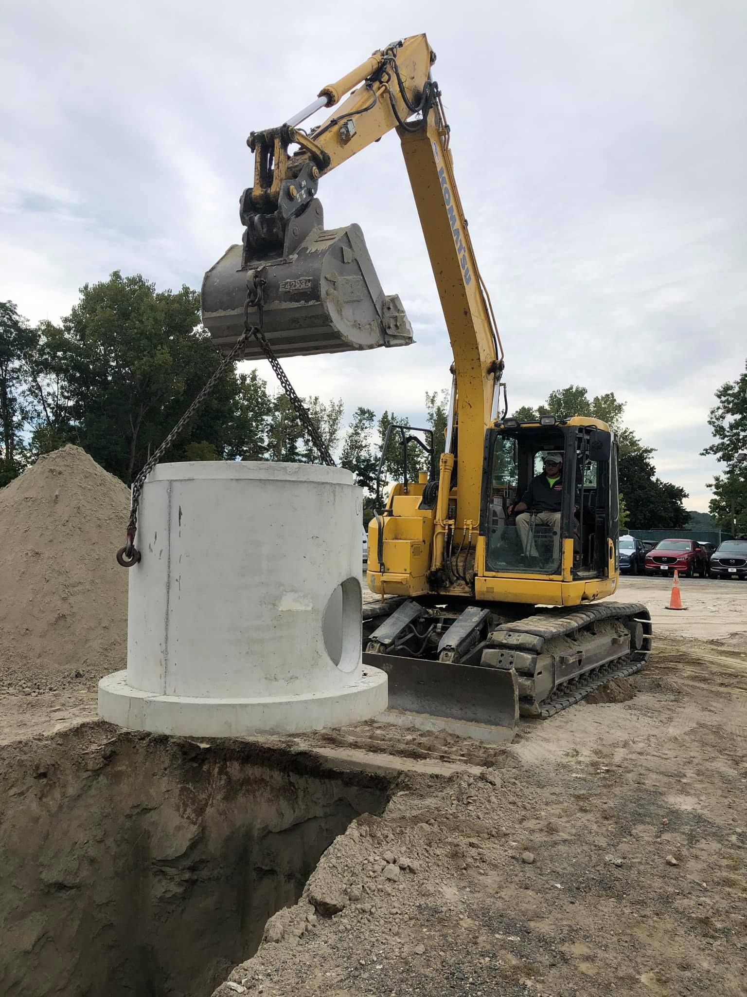 Yellow excavator lifting a concrete cylinder over a dug hole; construction site.