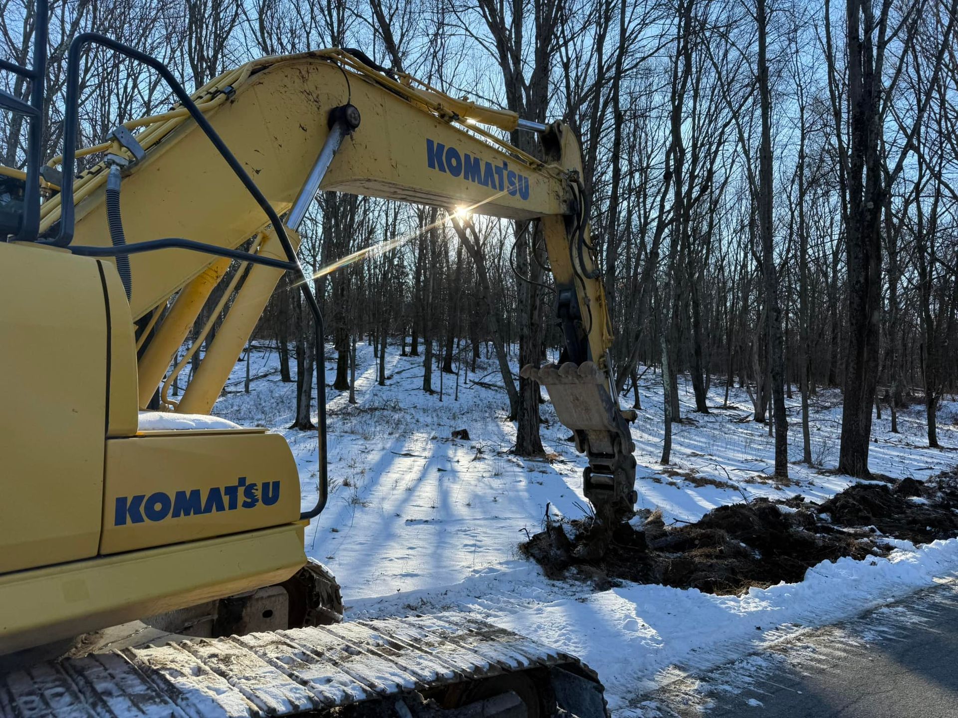 Yellow Komatsu excavator digging trench in snowy area near trees. Sunlight visible.