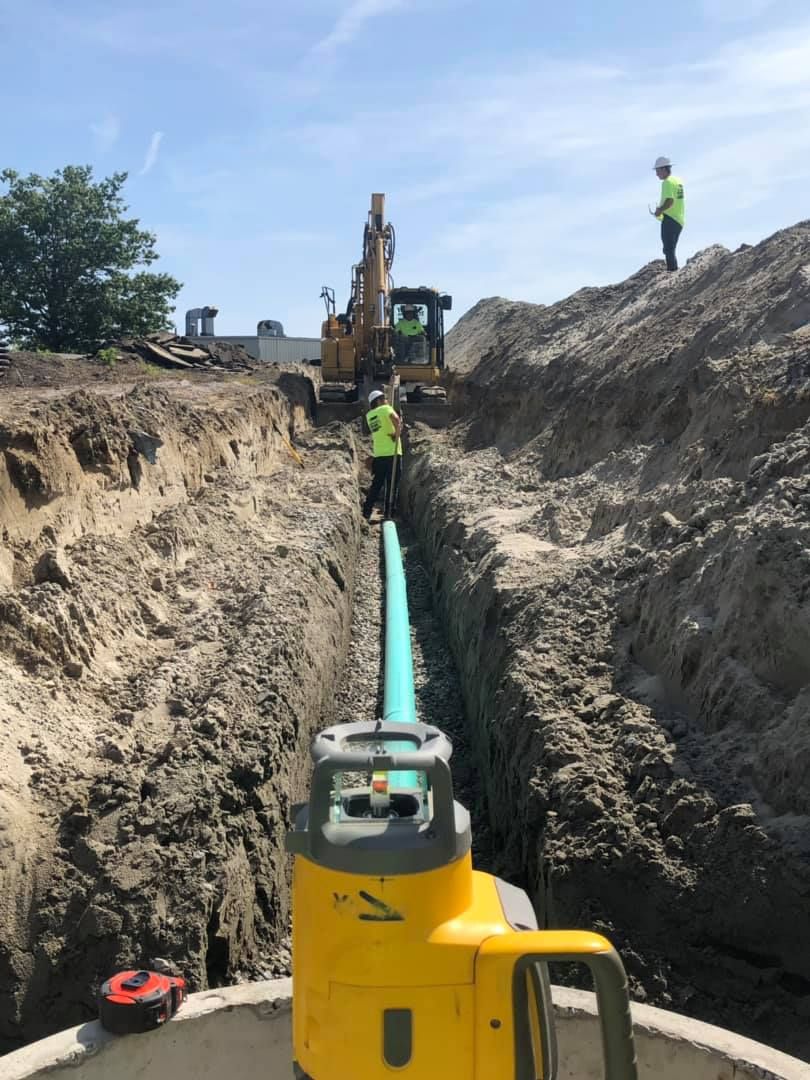 Construction crew working in a trench, installing pipe. An excavator, laser level, and workers in safety vests are visible.
