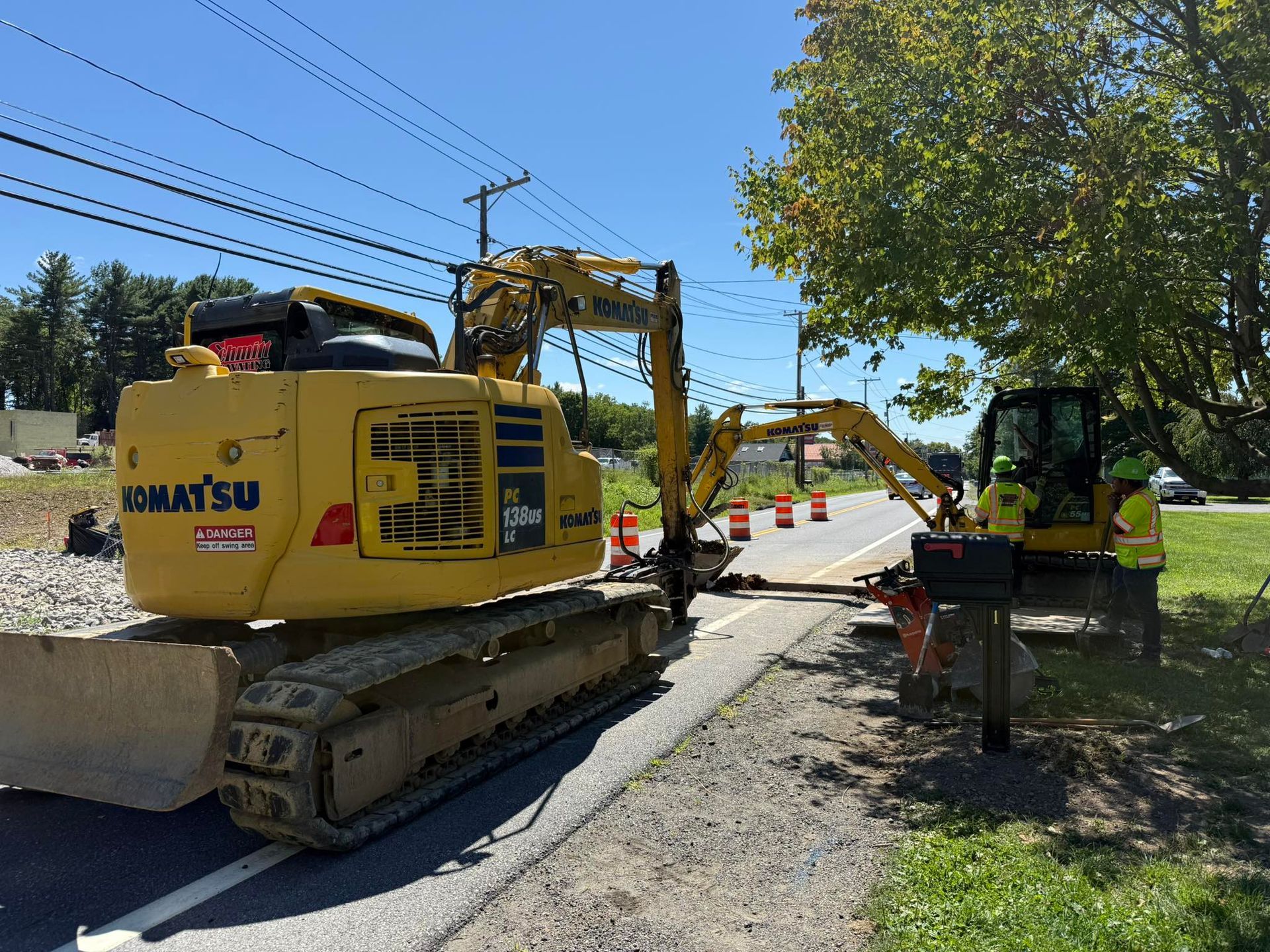 Two excavators working on road repairs next to a road with traffic cones. Sunny day.