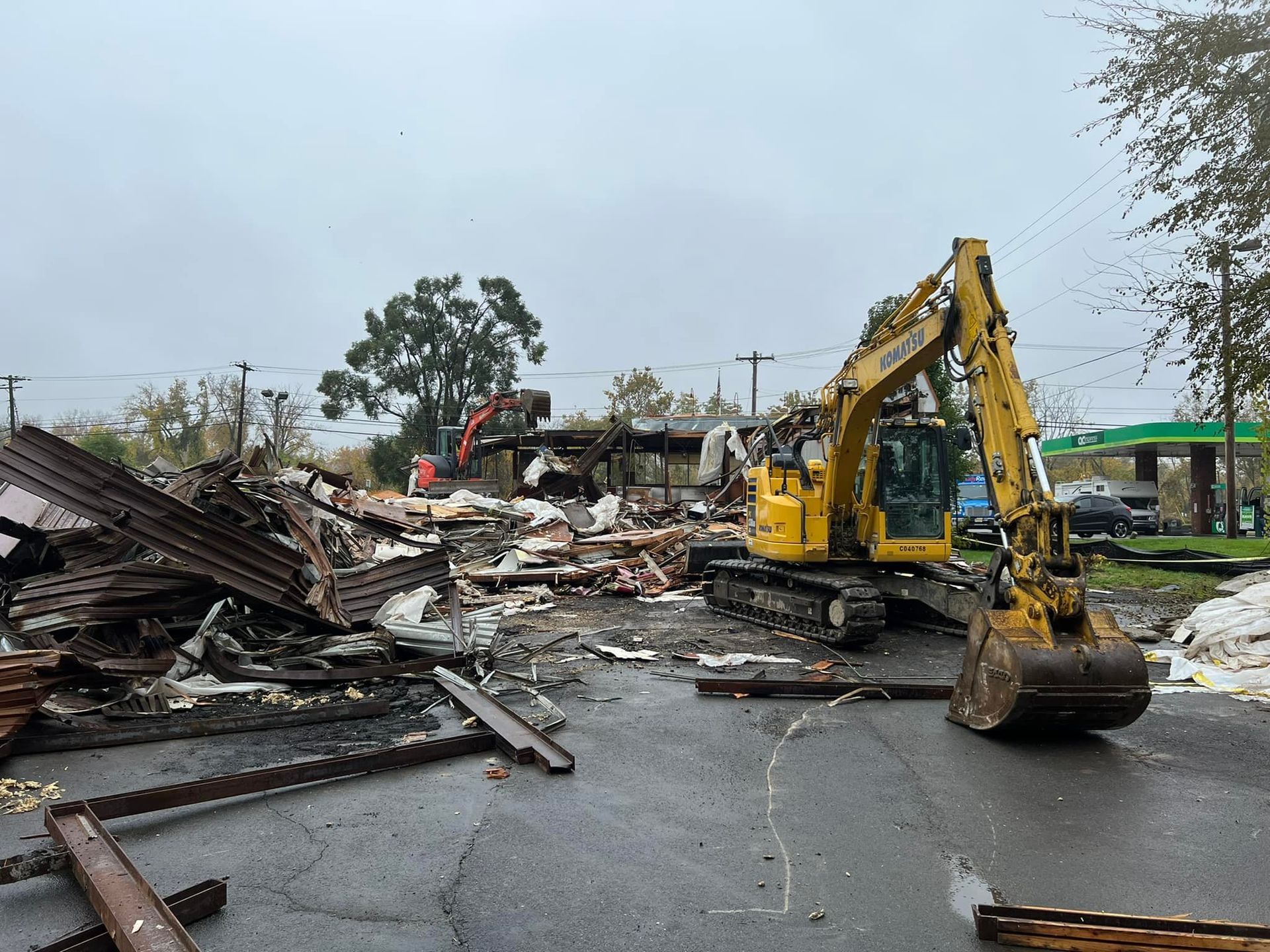 A yellow excavator demolishes a building on a gray, overcast day; debris litters the ground.
