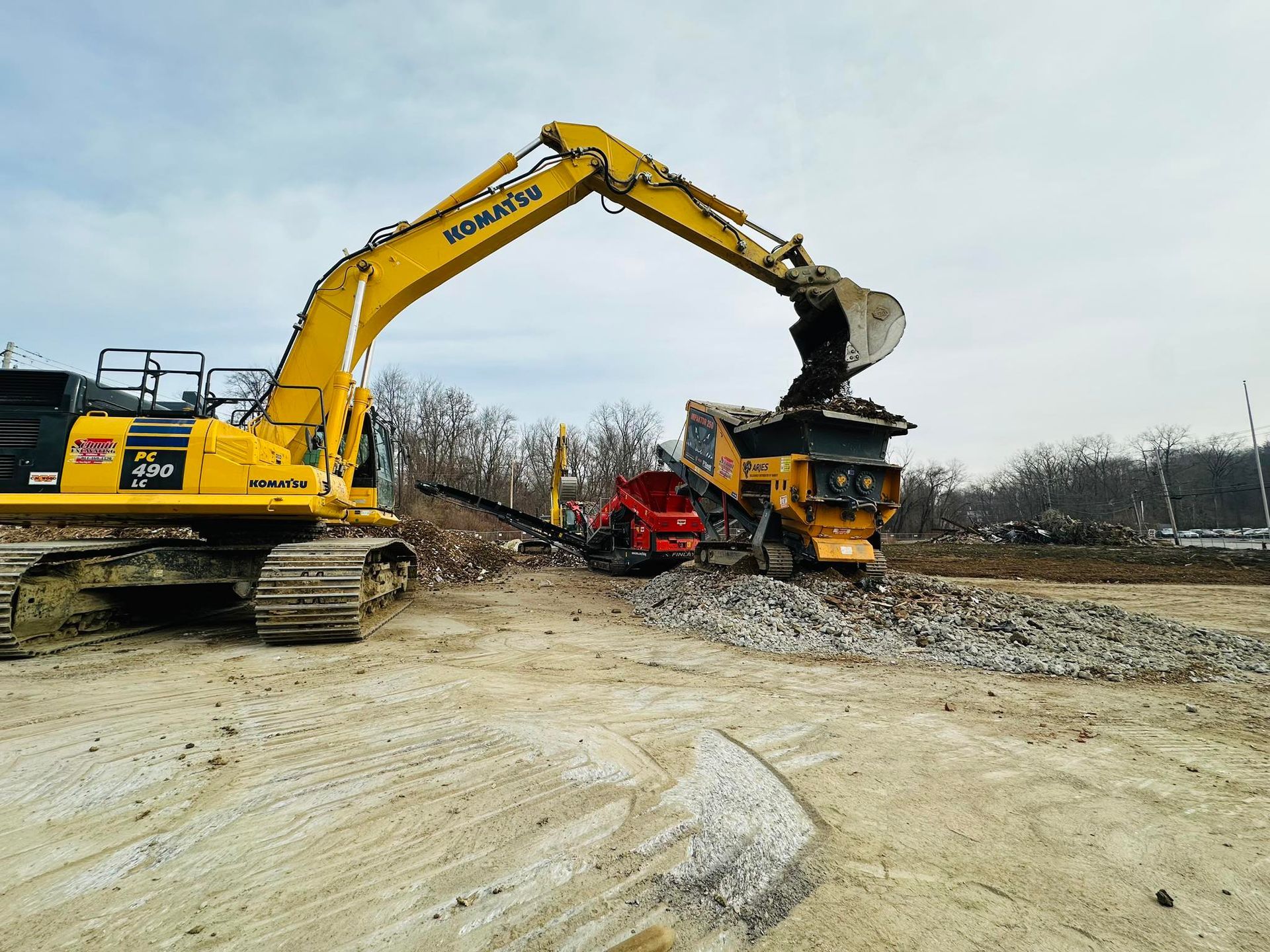 Yellow excavator dumping debris into a rock crusher on a construction site.