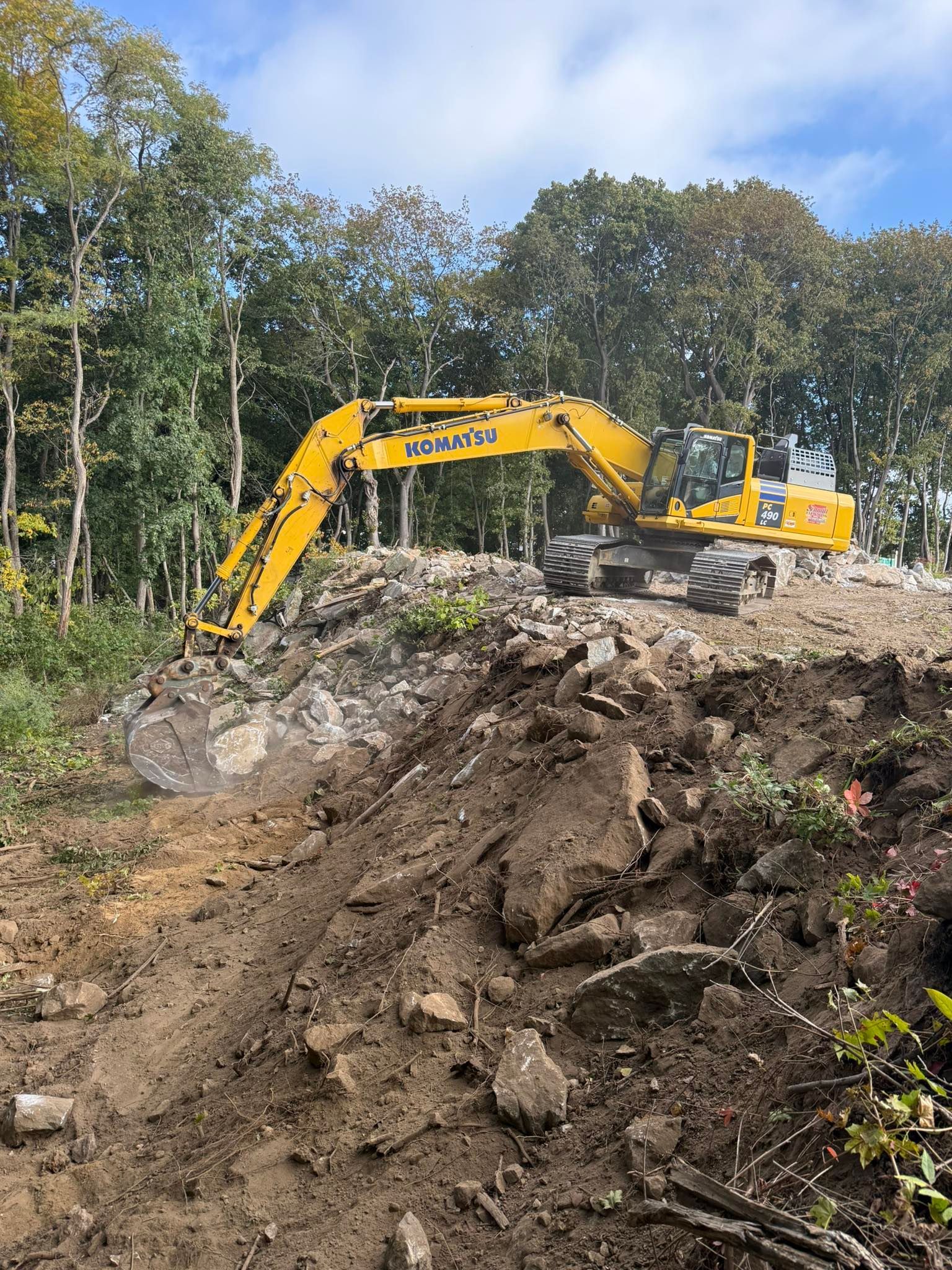 Yellow excavator on a rocky hill, clearing land near trees under a blue sky.