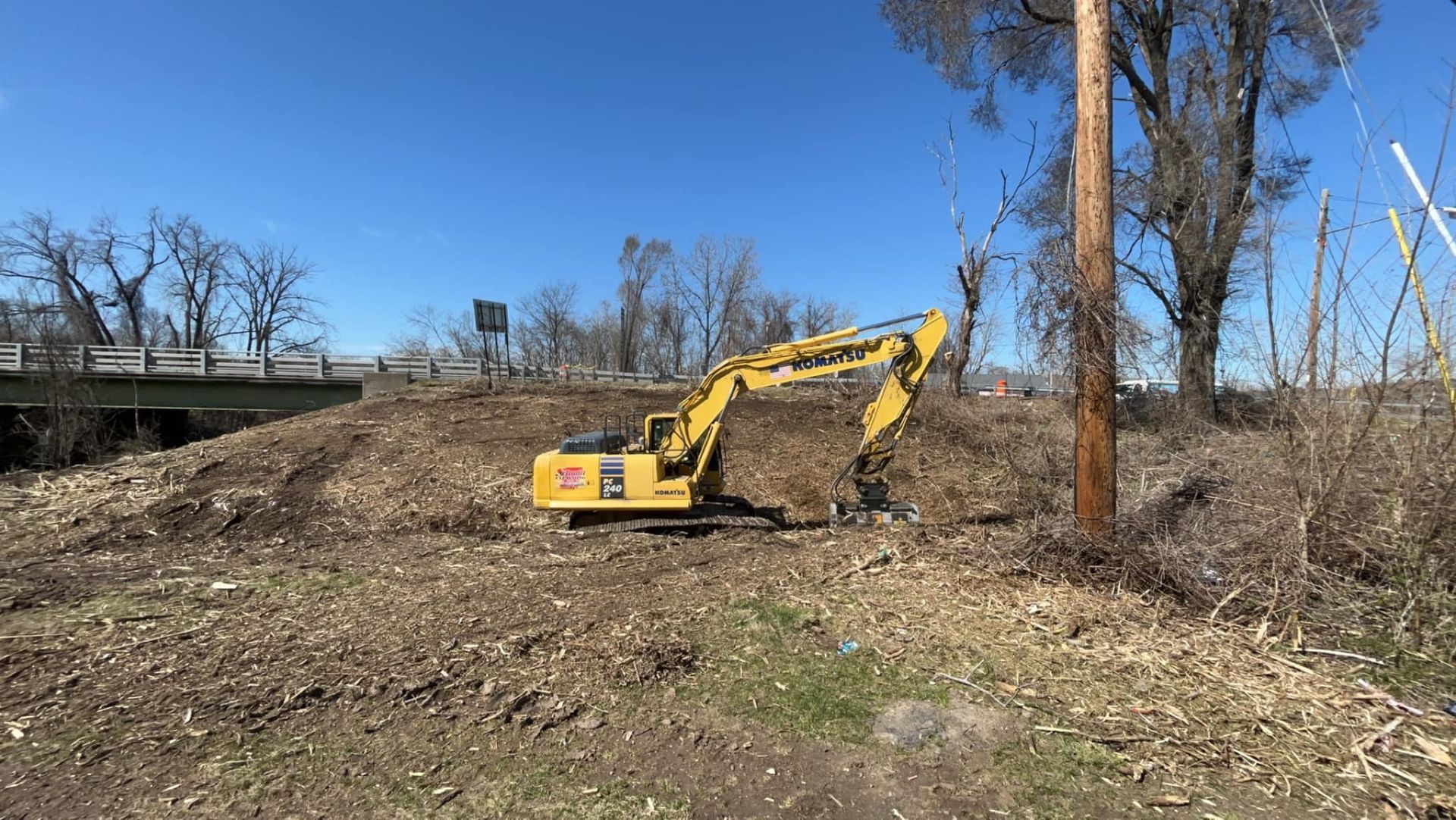 Yellow excavator on a pile of wood chips next to a utility pole under a blue sky.