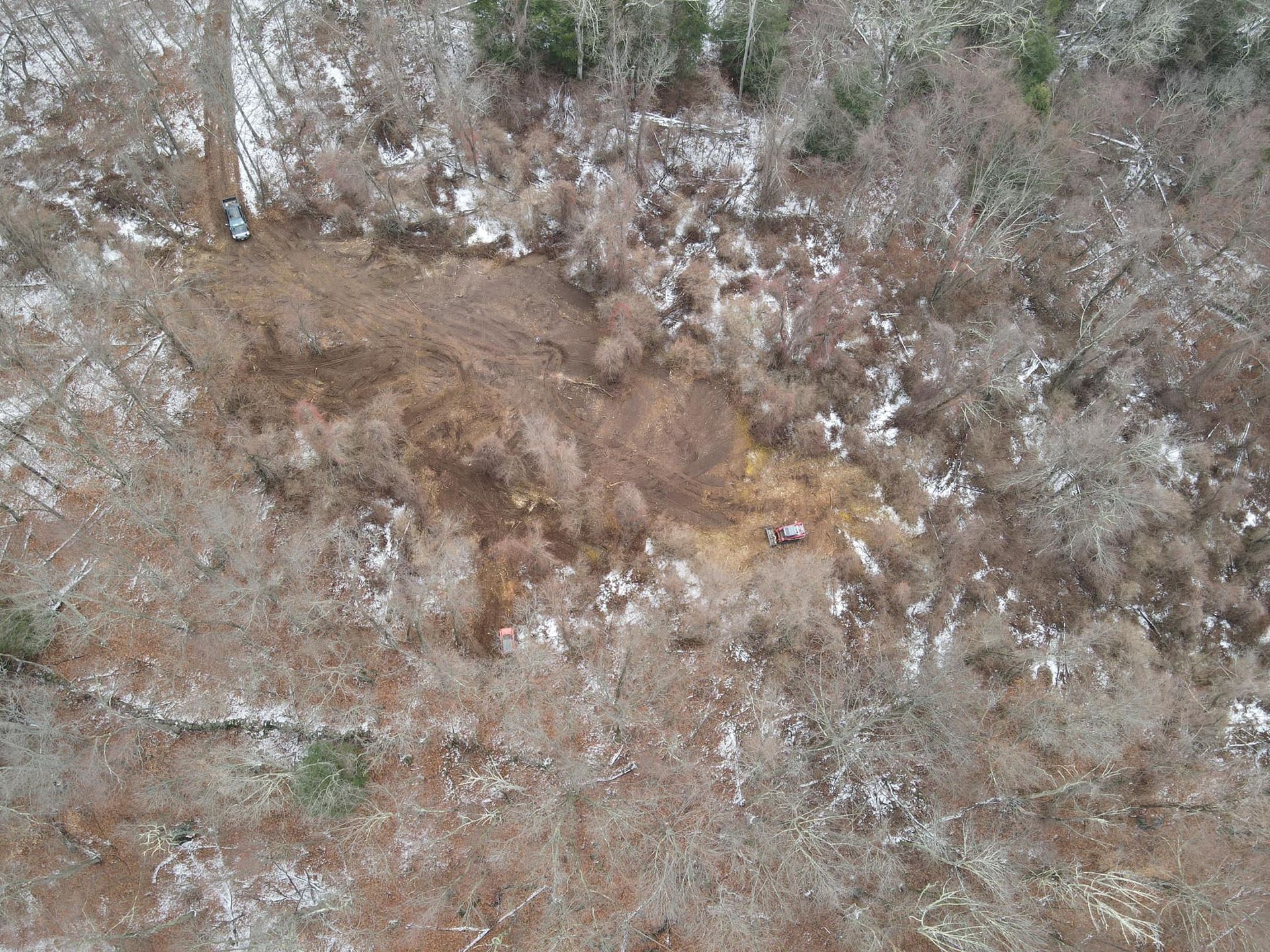 Aerial view of a snowy forest clearing with two vehicles and brown foliage, suggesting a recent activity.