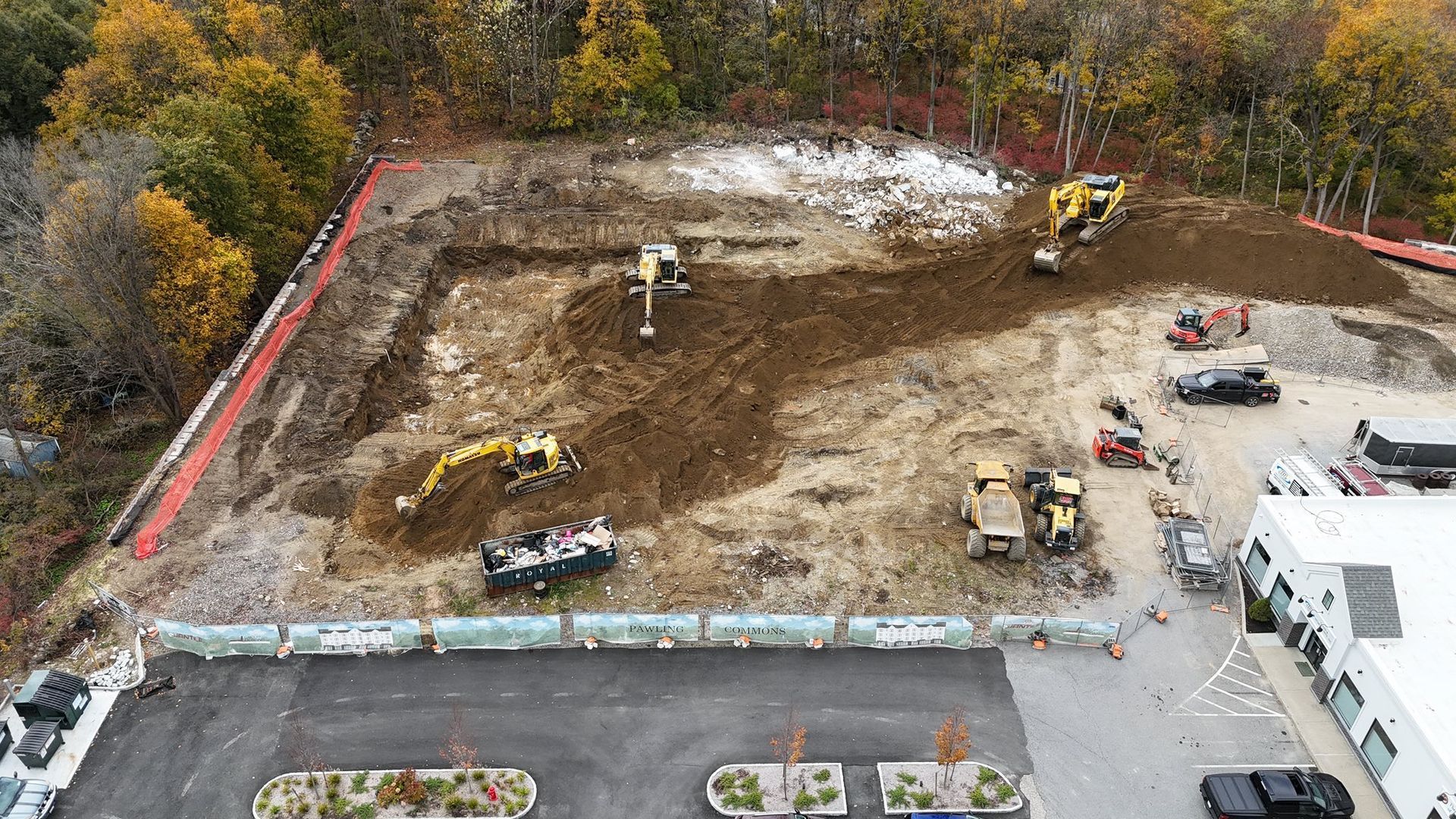 Construction site with several excavators and trucks, surrounded by trees and a retaining wall.