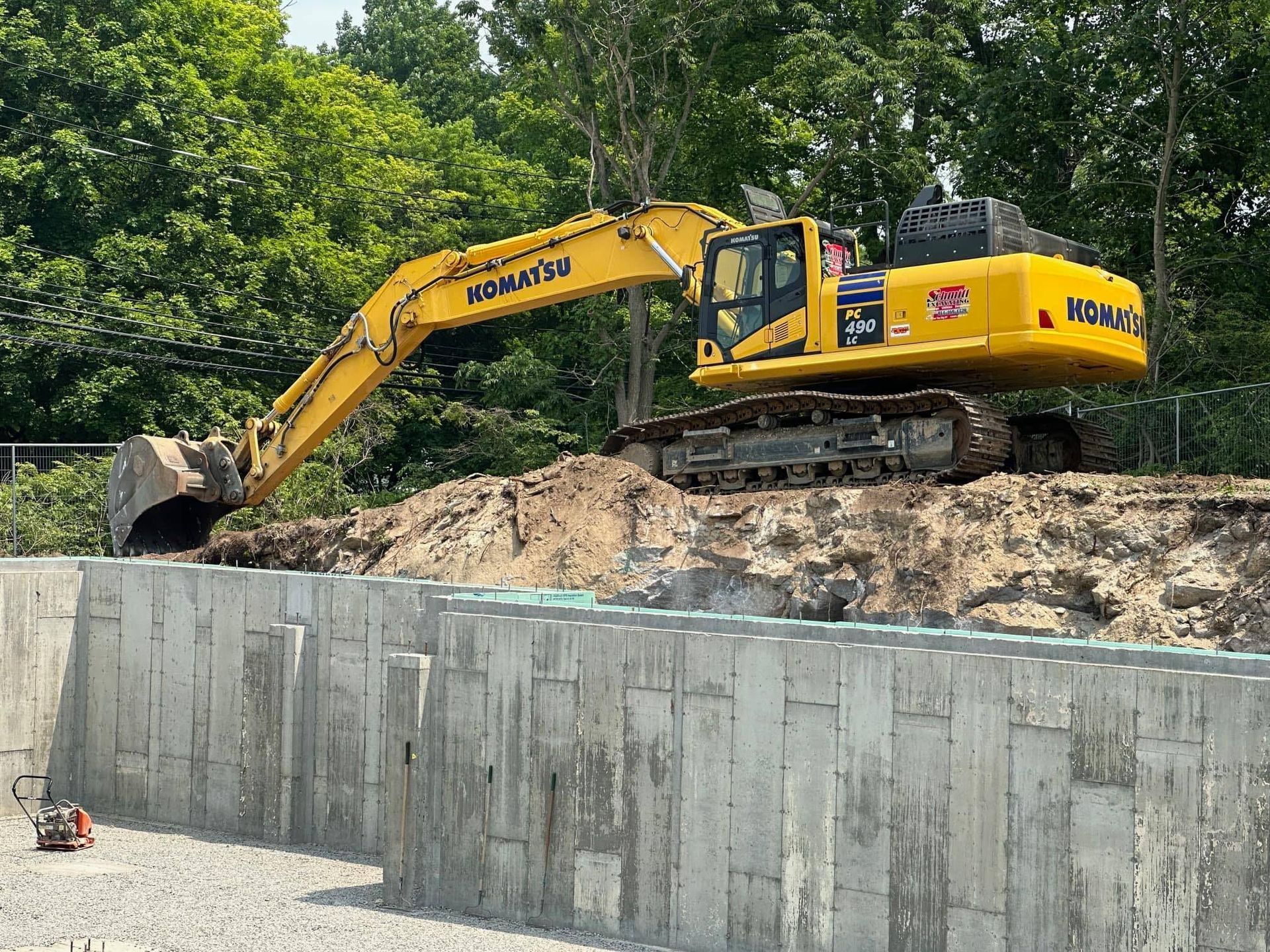 Yellow Komatsu excavator digging on a dirt mound, next to a concrete retaining wall.