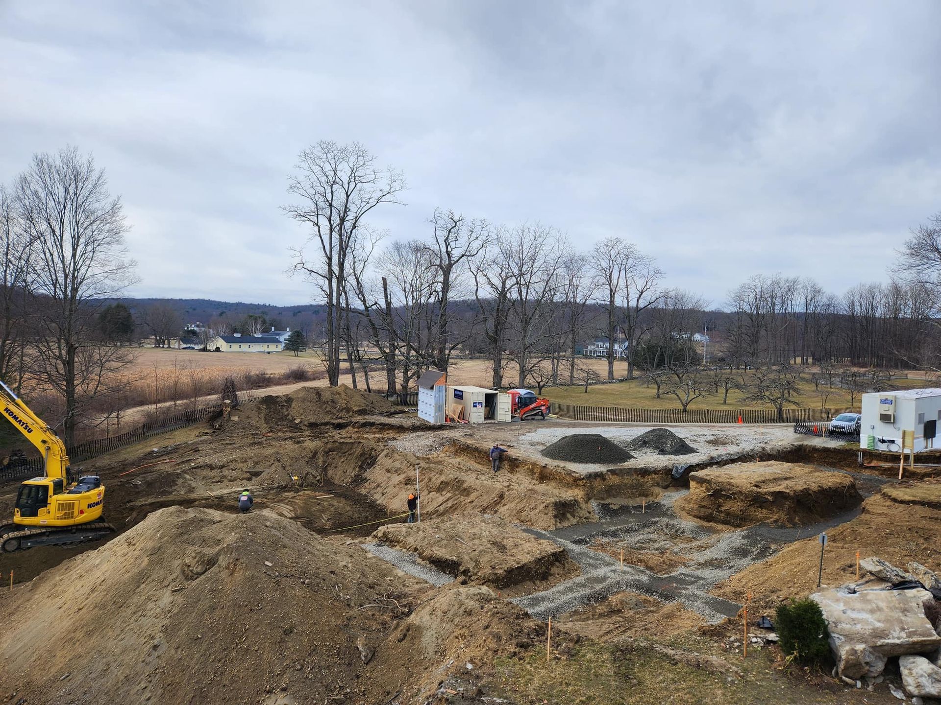 Construction site with excavator and dirt piles; cloudy sky, trees in background.