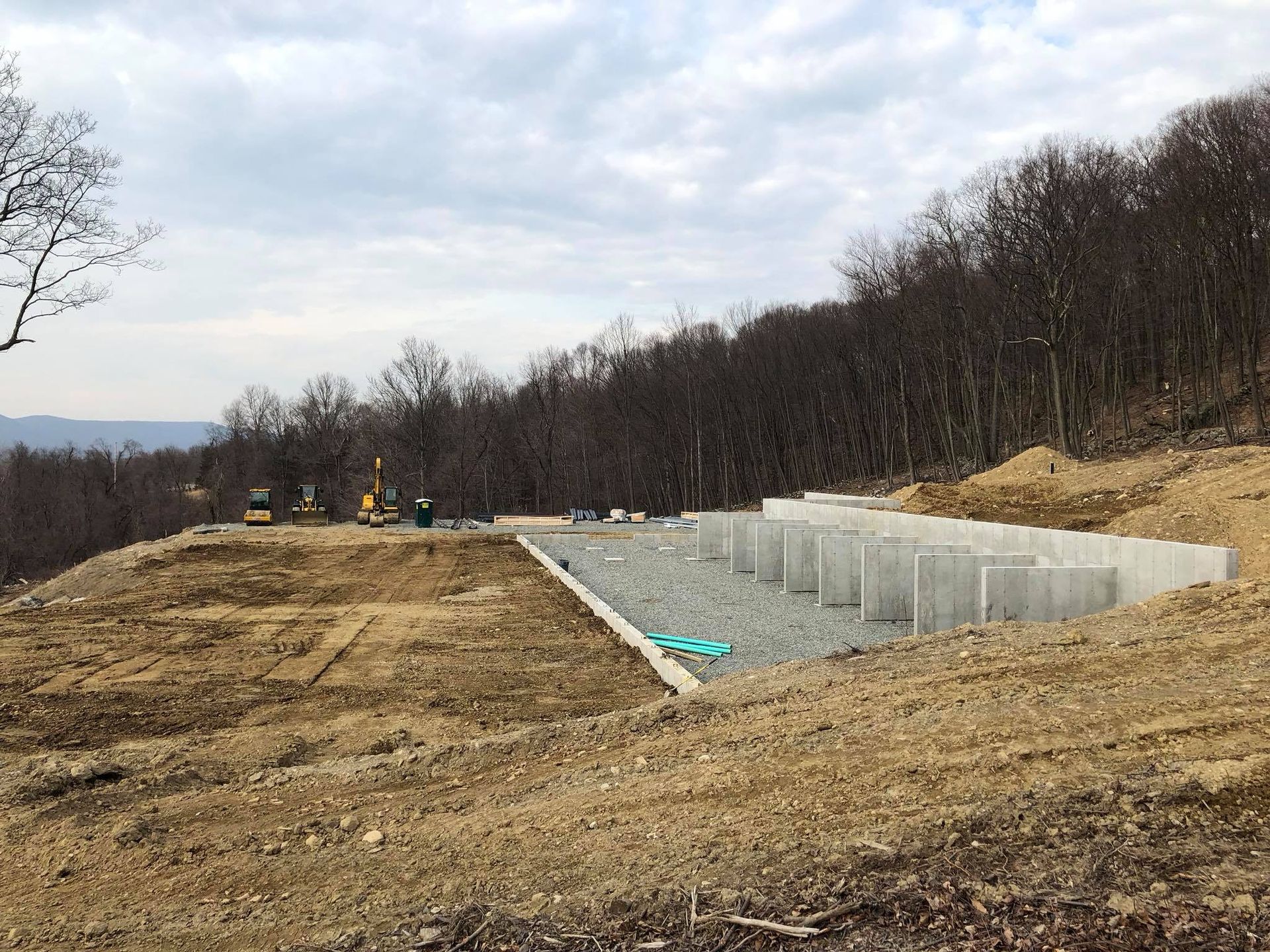 Construction site with concrete foundation walls on a hillside, surrounded by dirt and trees.