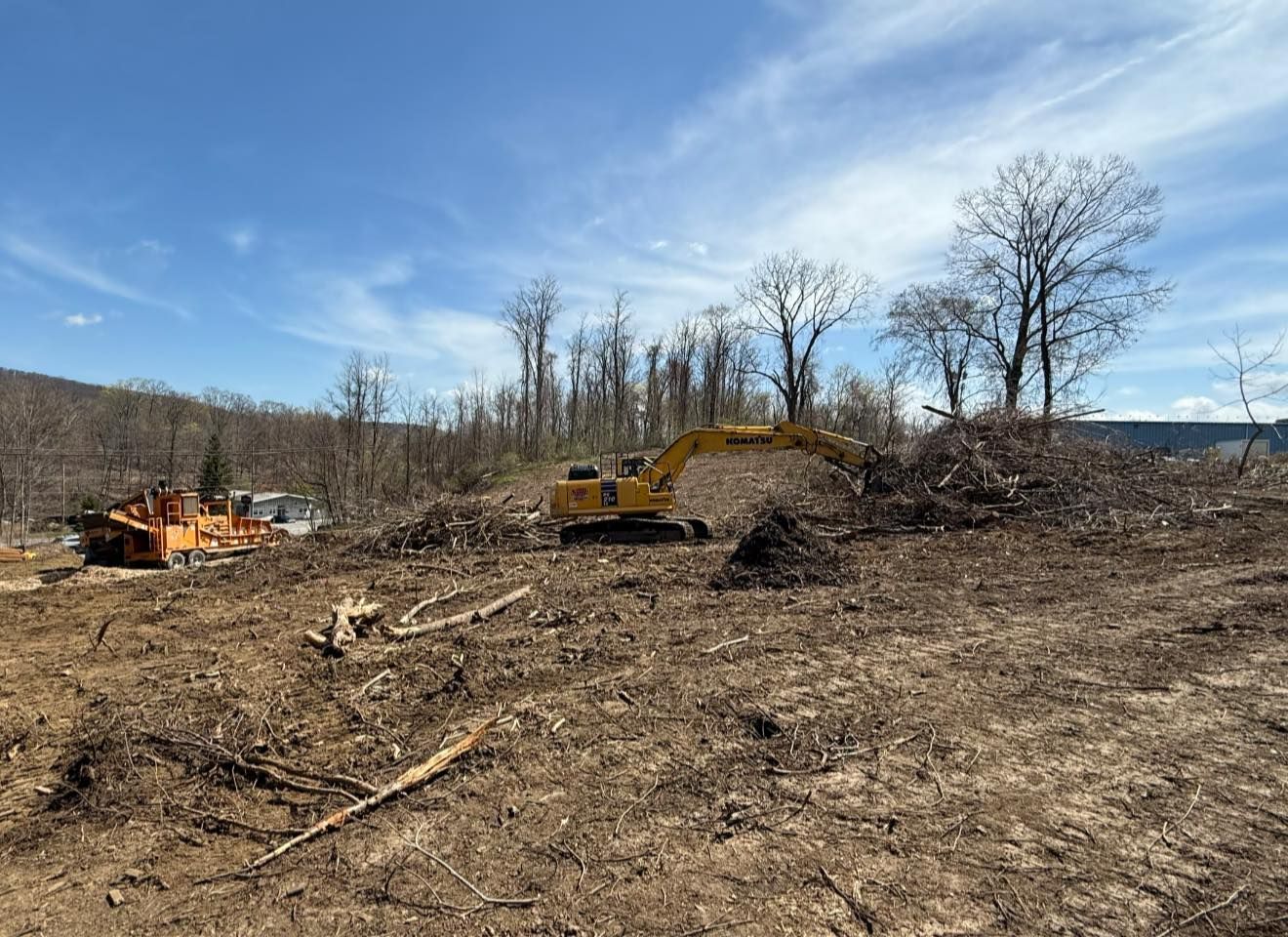 An excavator clears land, moving debris. Bare trees and blue sky background.